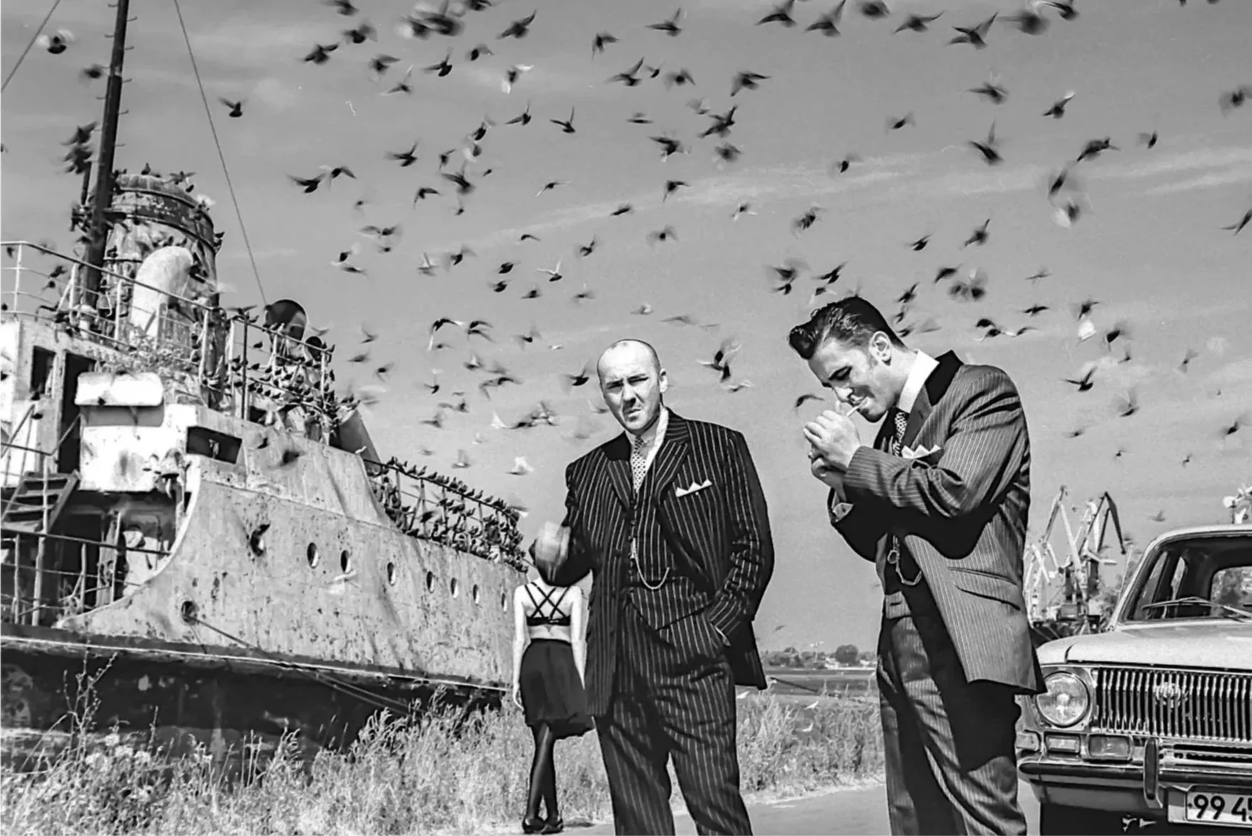 Black-and-white photo of two men in suits standing outdoors near a docked ship, with birds flying overhead. One man is gesturing with his fist, while the other is smoking a cigarette. A car is parked nearby.