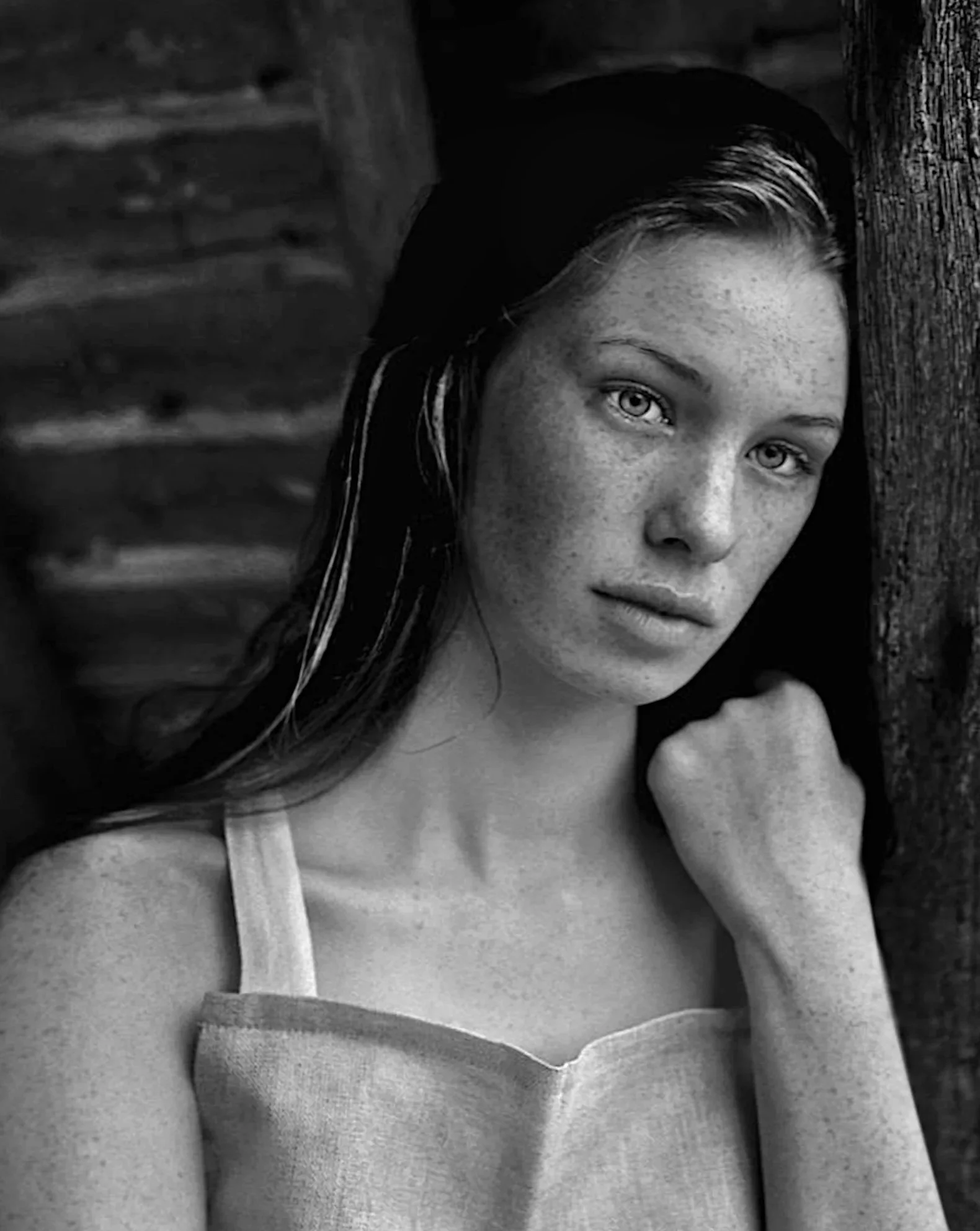 A black and white portrait of a young woman with light skin and freckles, looking at the camera, leaning against a wooden post, with a wooden background.