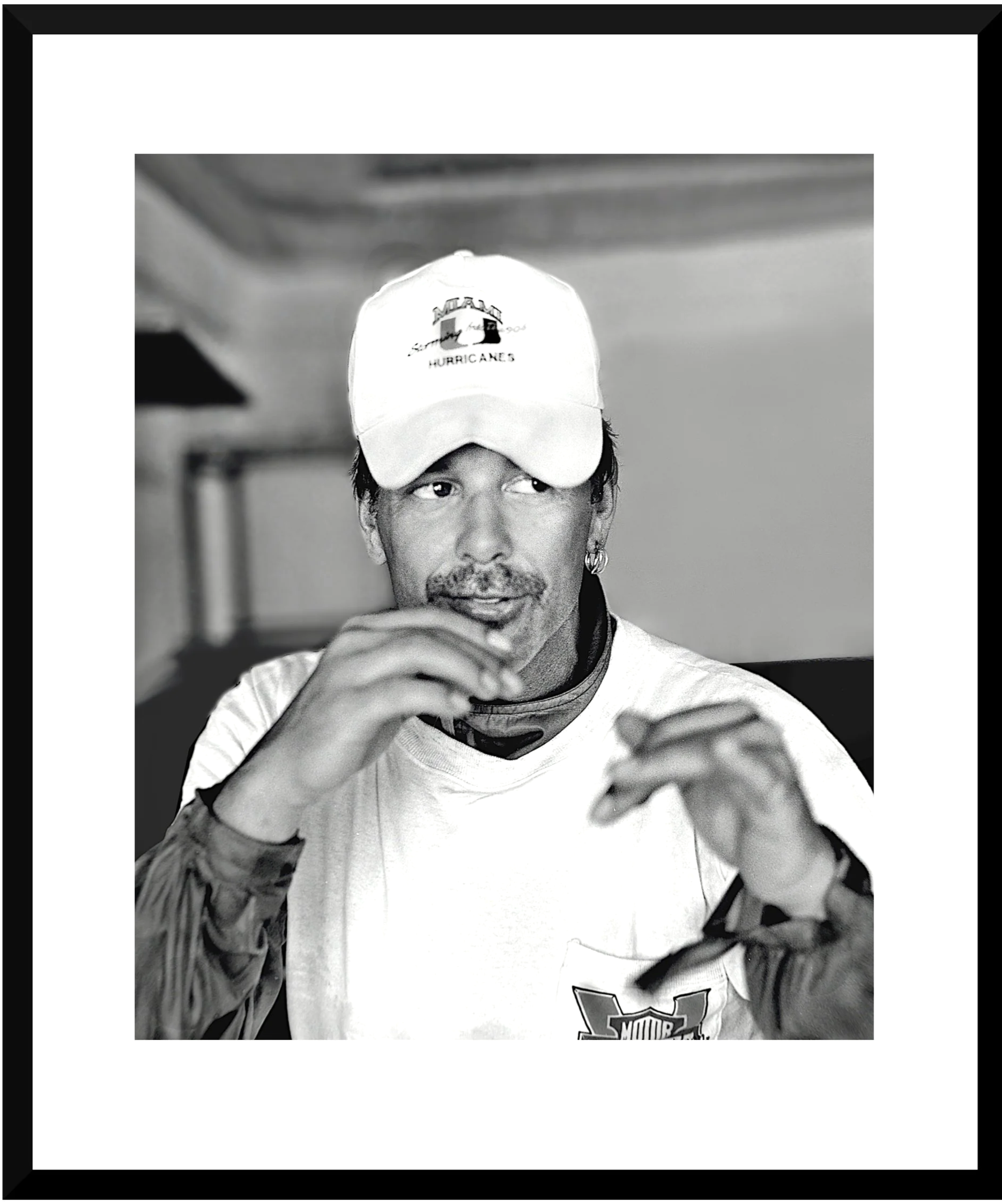 Black and white photo of a man wearing a Miami Hurricanes cap and a white T-shirt, with tattoos on his arm. He is smiling slightly and is holding a pair of glasses in his hand, looking to the side.