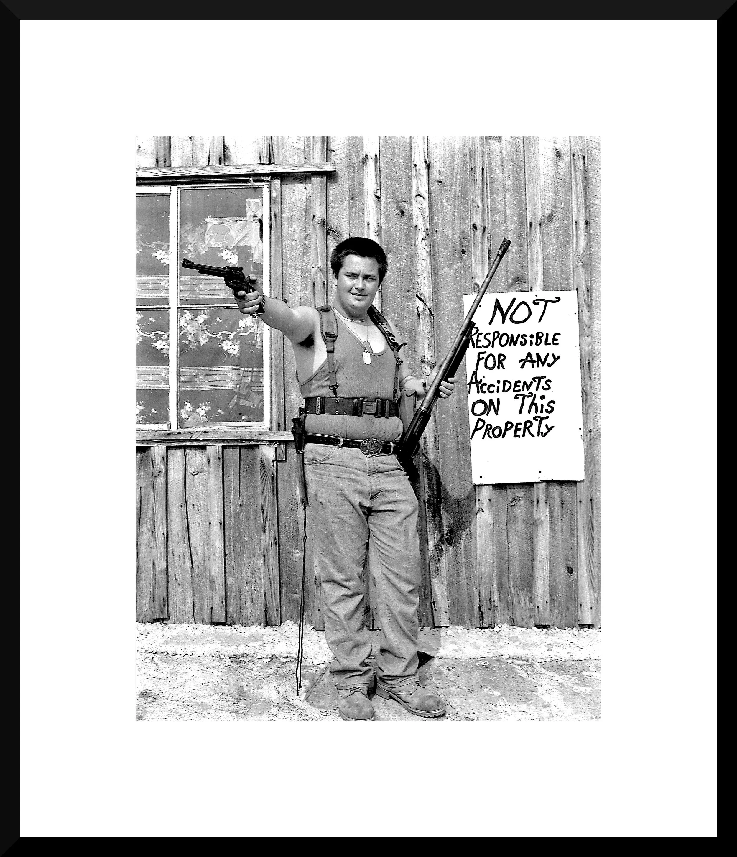 A person standing outdoors in front of a wooden building with a window, holding a pistol in one hand and a rifle in the other, wearing tactical gear, and a sign on the wall that reads 'Not responsible for any accidents on this property.'