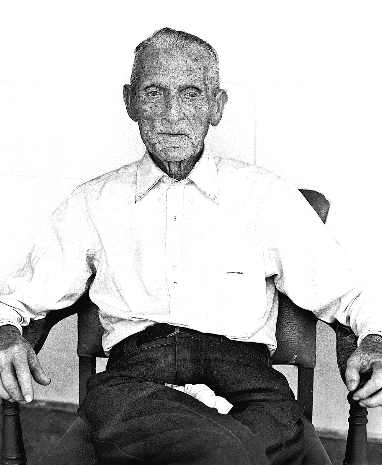 An elderly man with gray hair and a serious expression, wearing a light-colored button-up shirt, seated in a chair against a plain background.