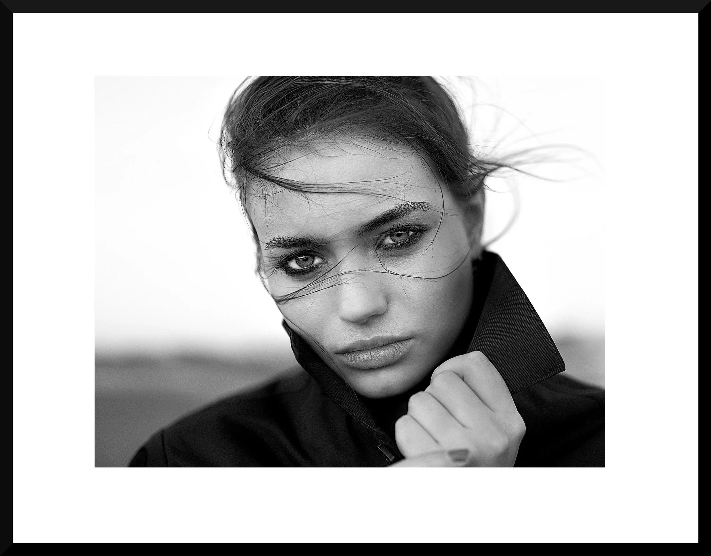 Black and white close-up portrait of a woman with wind-blown hair, intense gaze, and slightly parted lips, wearing a high-collared jacket.
