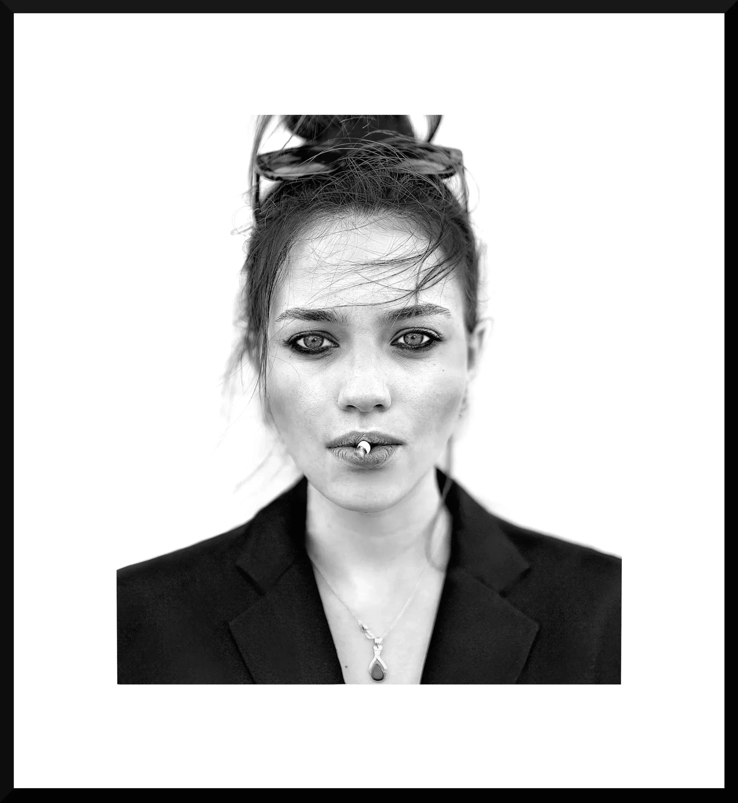 Black and white portrait of a woman with light skin, wearing a blazer and a necklace, with her hair tied up and sunglasses on her head. She has a cigarette in her mouth and a confident expression.