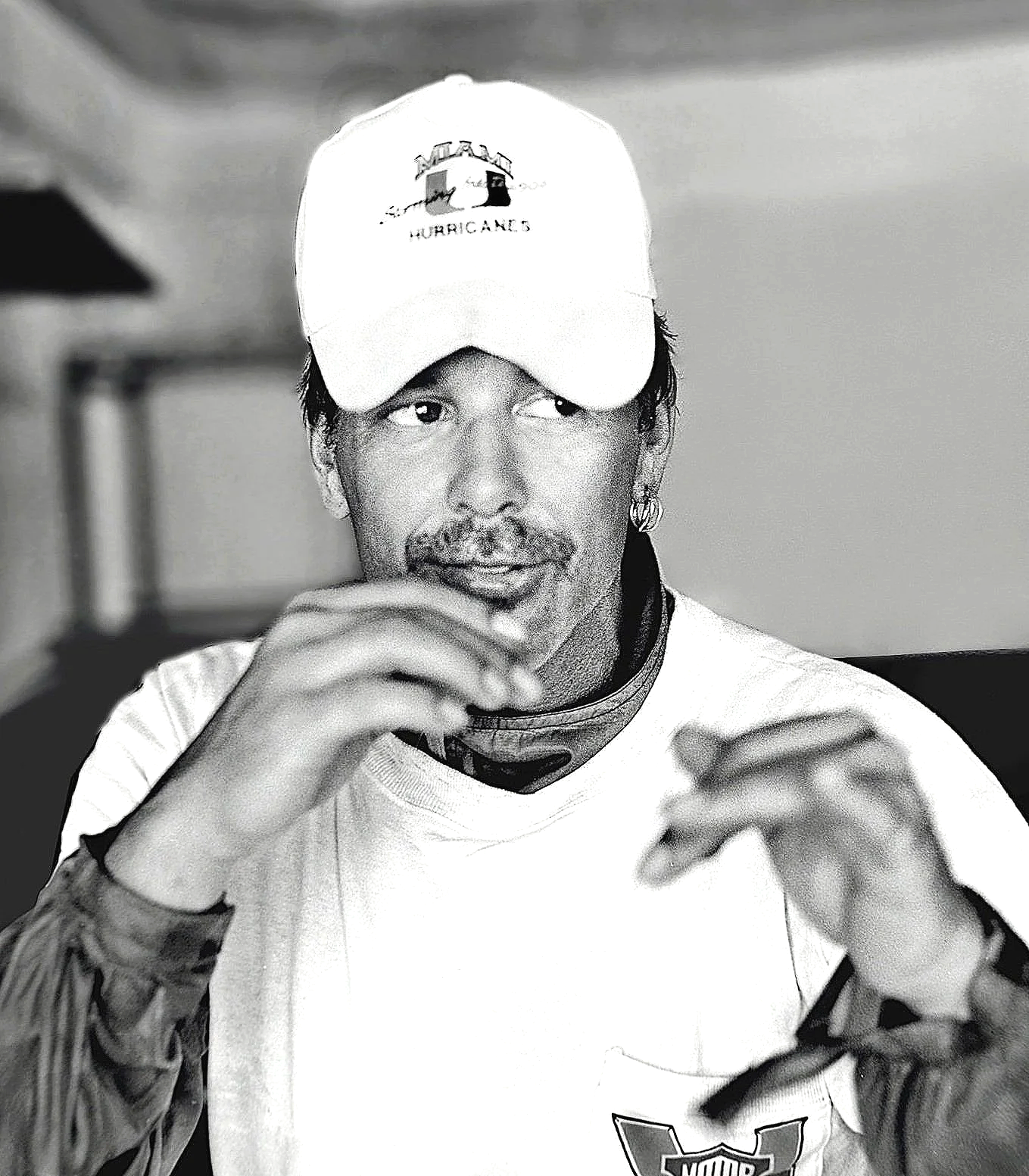 Black and white photo of a man wearing a baseball cap and a racing suit, adjusting his sunglasses or glasses, with tattoos visible on his arms.