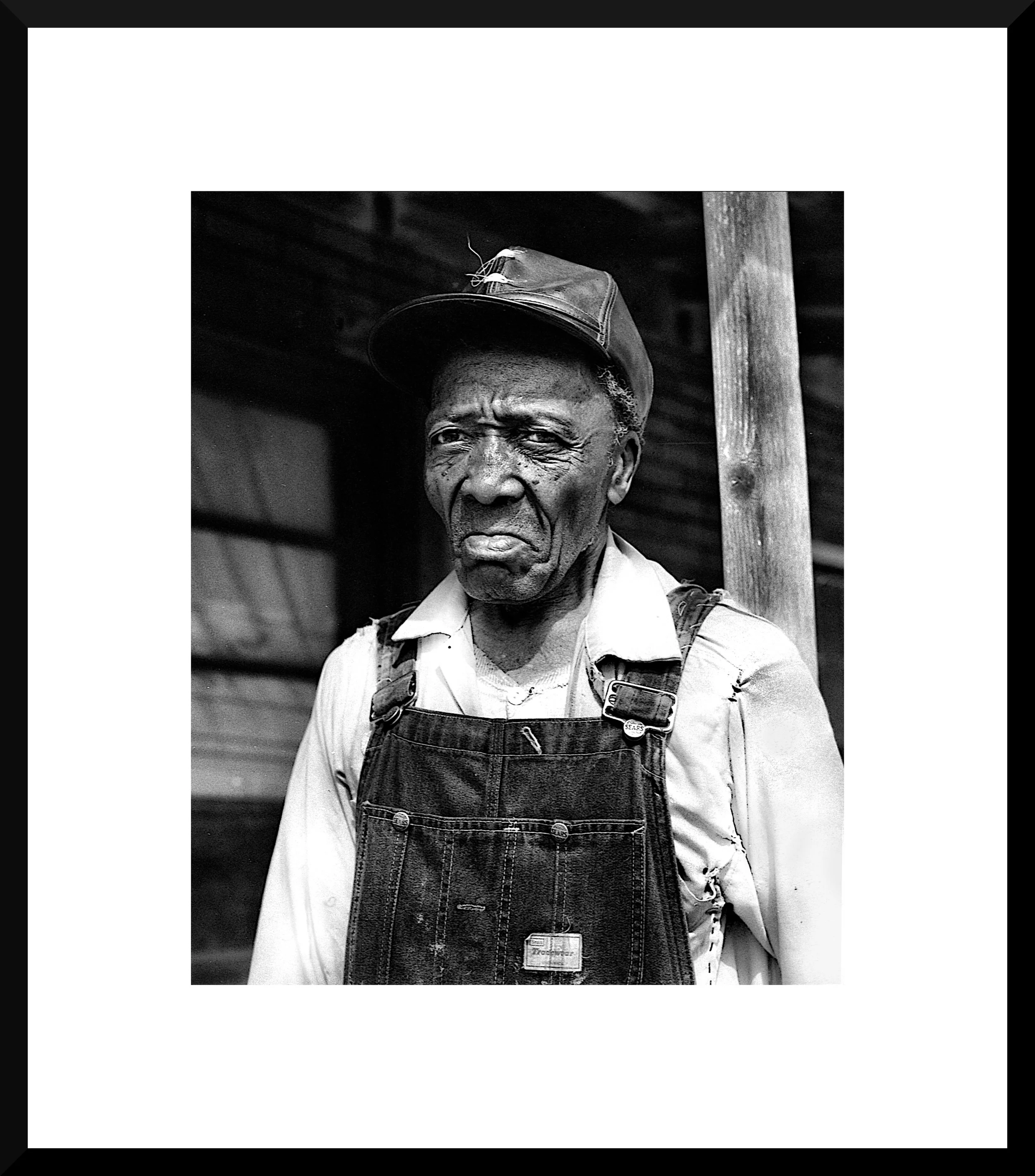 Black and white photo of an elderly man wearing a cap, overalls, and a button-up shirt, standing outdoors near a wooden post.