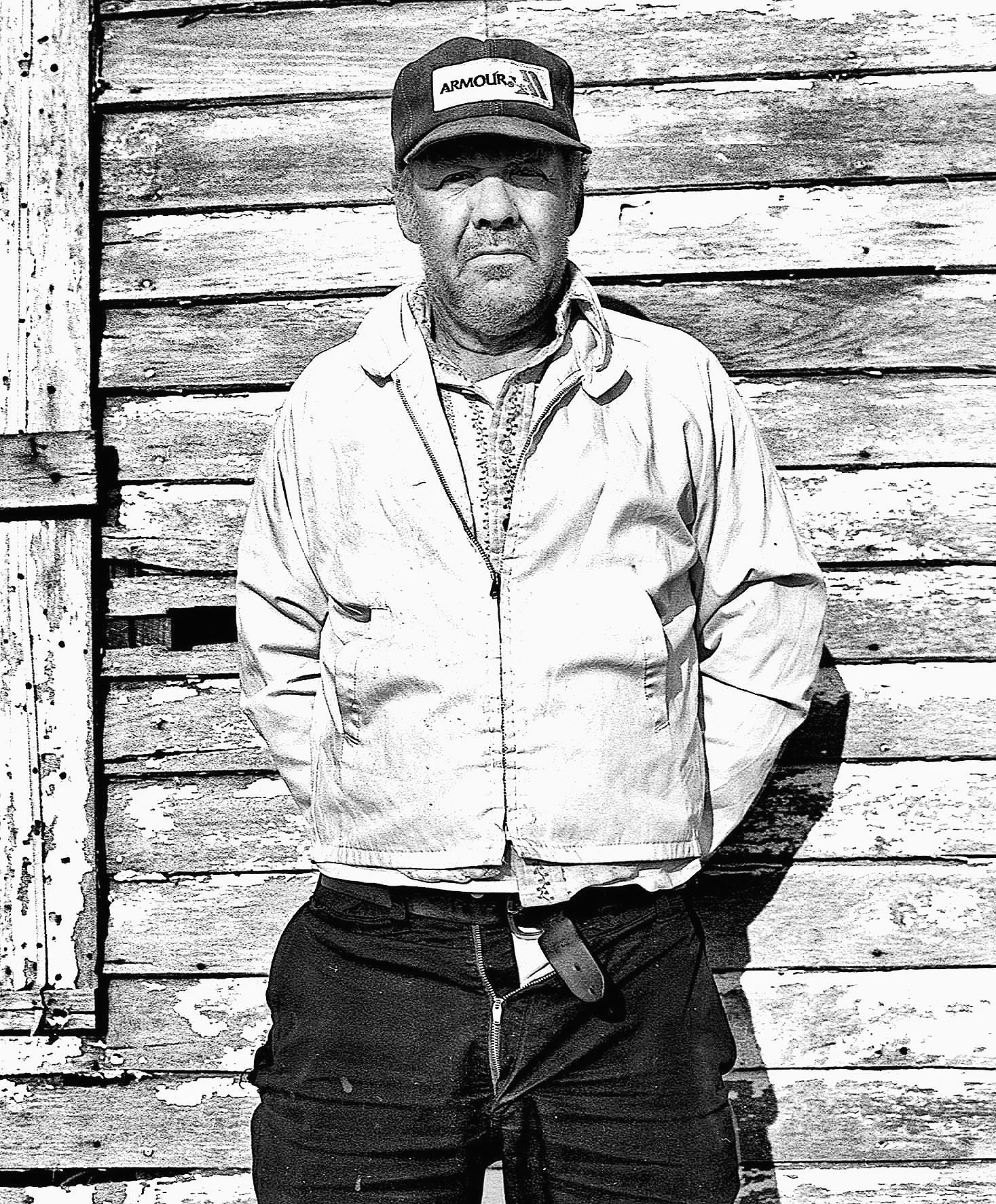 A black and white photo of a man standing in front of a weathered wooden wall, wearing a cap, a zip-up jacket, and dark pants.
