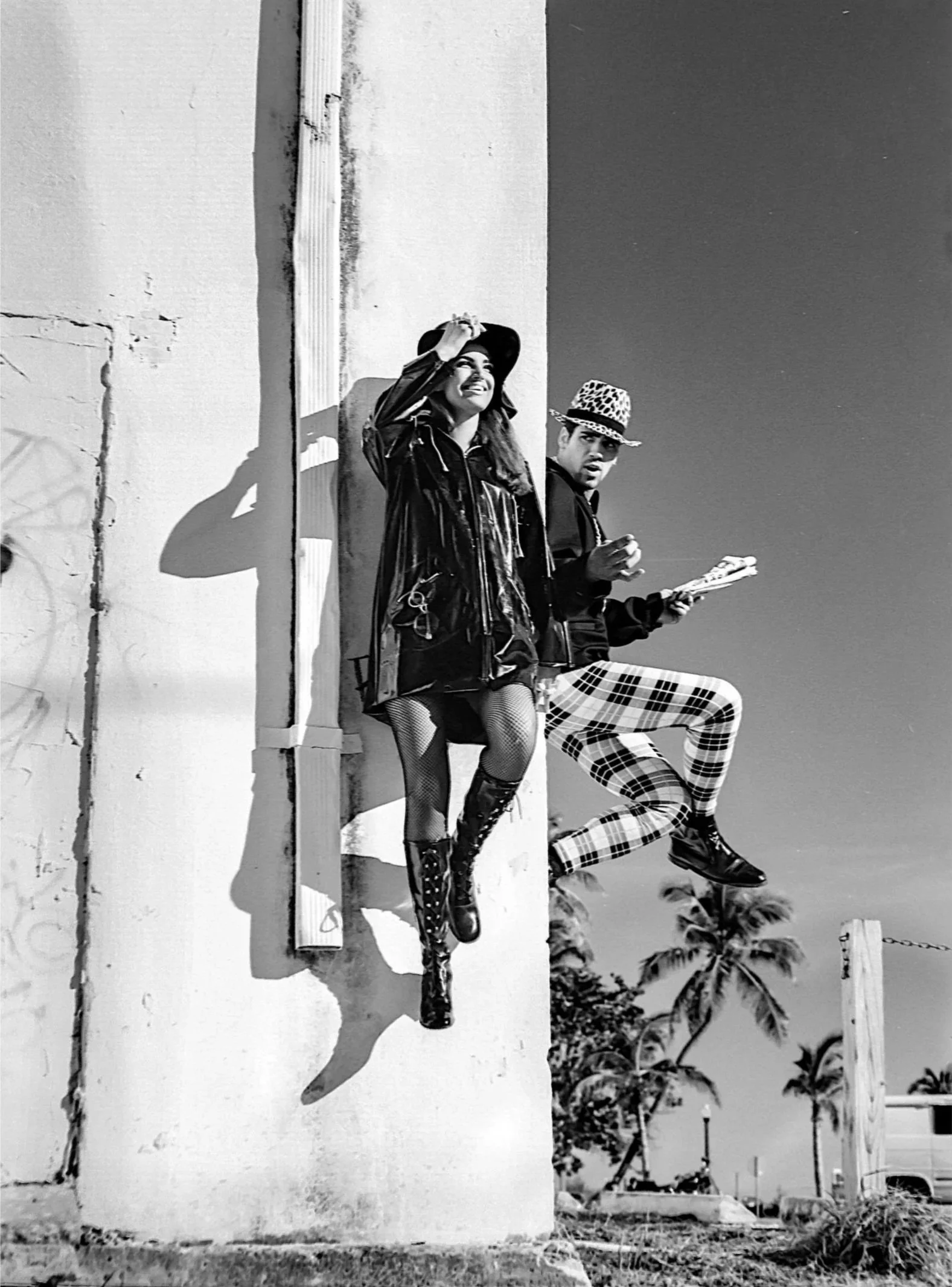 Black and white photo of two people sitting on a low wall, with a clear sky and trees in the background. The woman on the left is smiling, wearing a jacket, fishnet stockings, and knee-high boots. The man on the right is wearing a leopard-print hat, a dark jacket, plaid pants, and is holding a folded newspaper, with a surprised or curious expression.