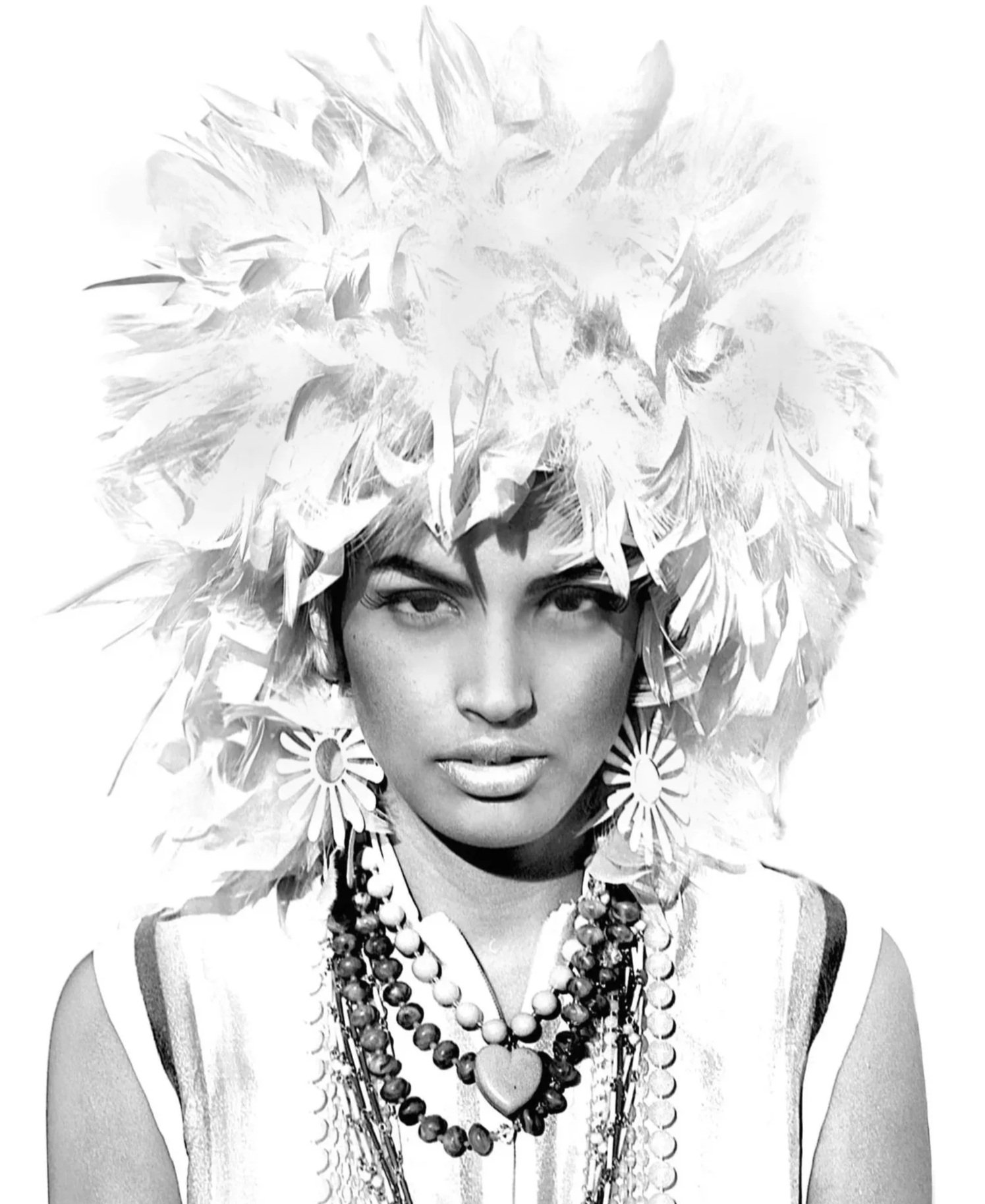 Black and white photo of a woman wearing a large, fluffy headdress with paper-like texture, floral earrings, layered necklaces, and a patterned top.