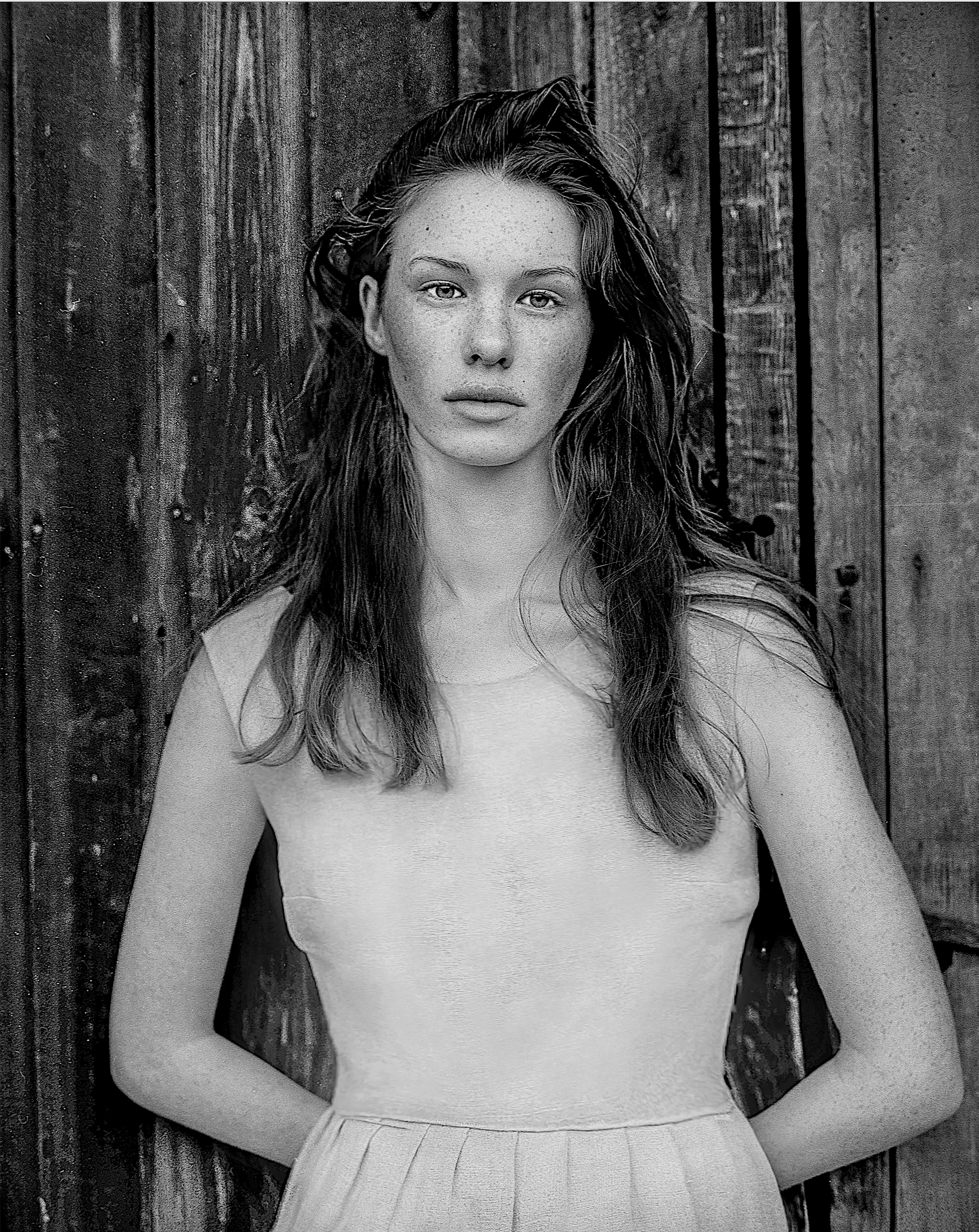 Black and white photo of a young woman with long wavy hair standing against a wooden background, wearing a sleeveless dress, looking directly at the camera.
