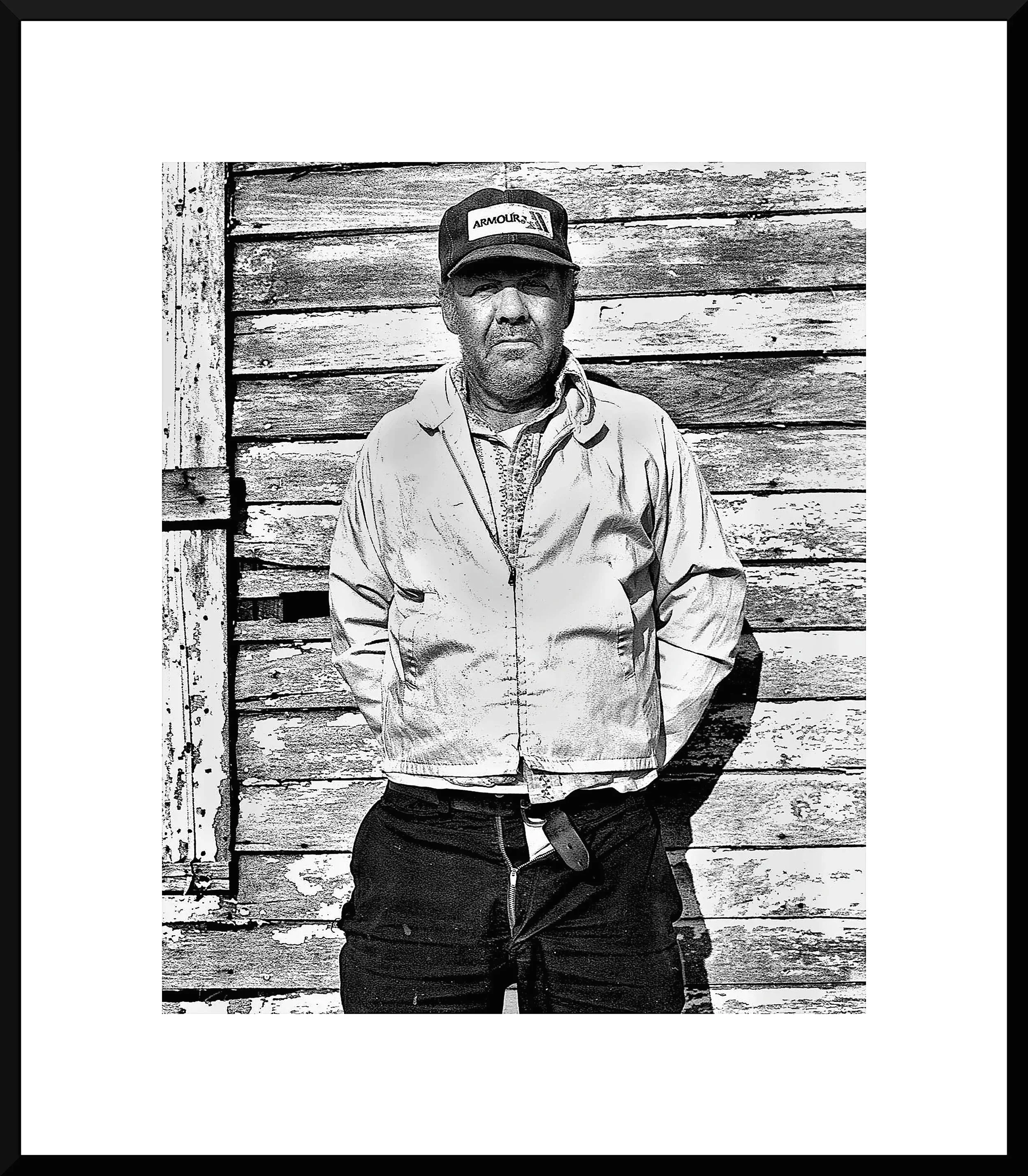 A man standing outdoors in front of a weathered wooden wall, wearing a cap, a light jacket, and dark pants.