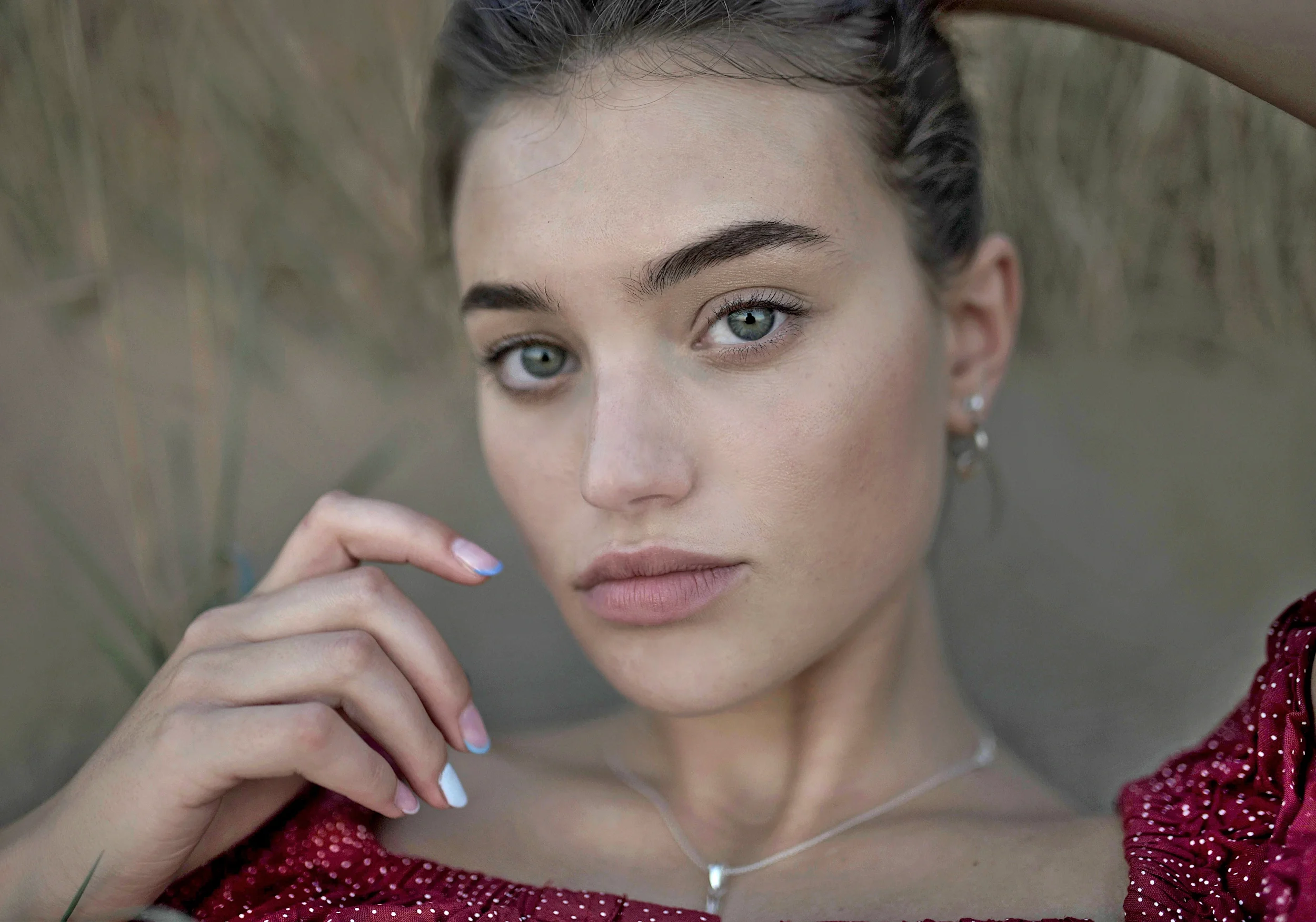 A close-up of a young woman with grayish-green eyes, full lips, and neatly groomed dark eyebrows. She has her hand near her face, wearing light pink lipstick and a silver necklace. She is dressed in a red polka-dot top with a slight V-neck. The background is blurred, showcasing a neutral, earthy tone.