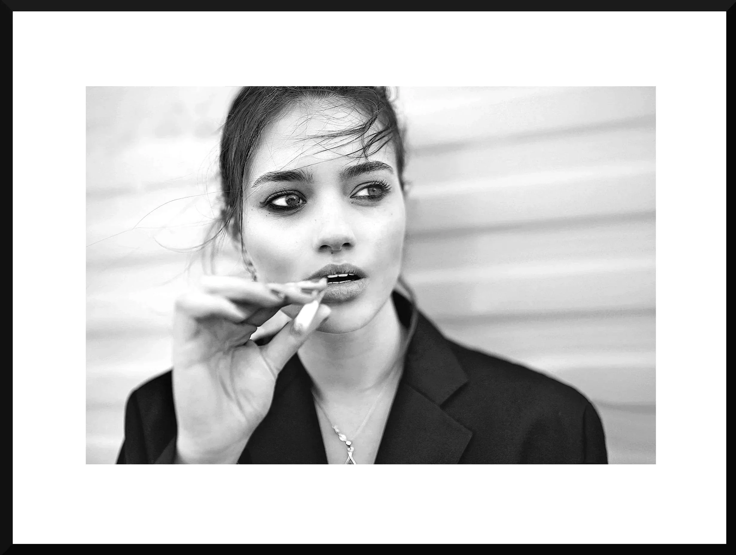 Black and white photo of a woman with dark hair and makeup, holding a cigarette near her lips, wearing a dark blazer and necklace, with a blurred background.