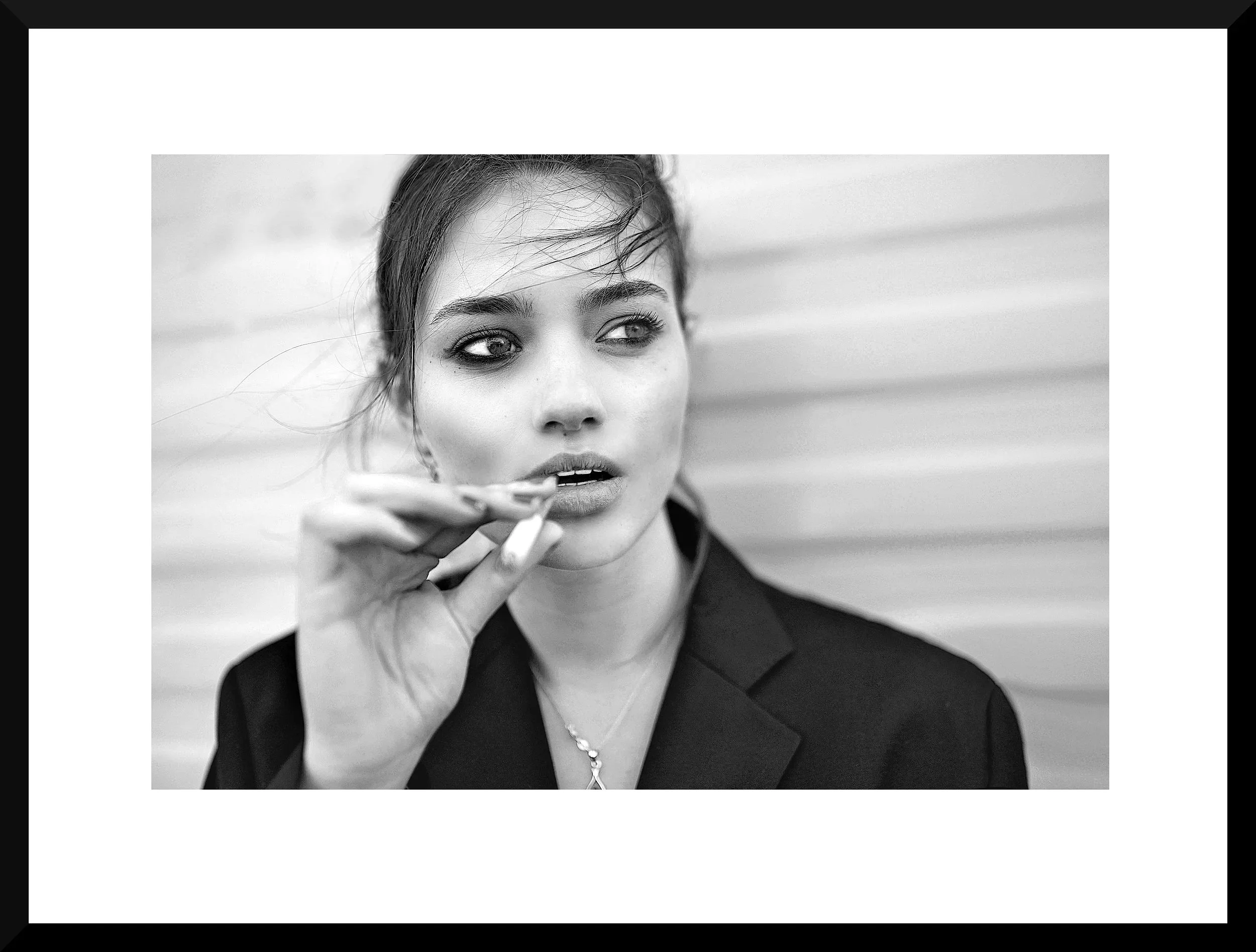 Black and white photograph of a woman with styled hair, wearing a blazer, and holding a cigarette near her lips, looking to the side.