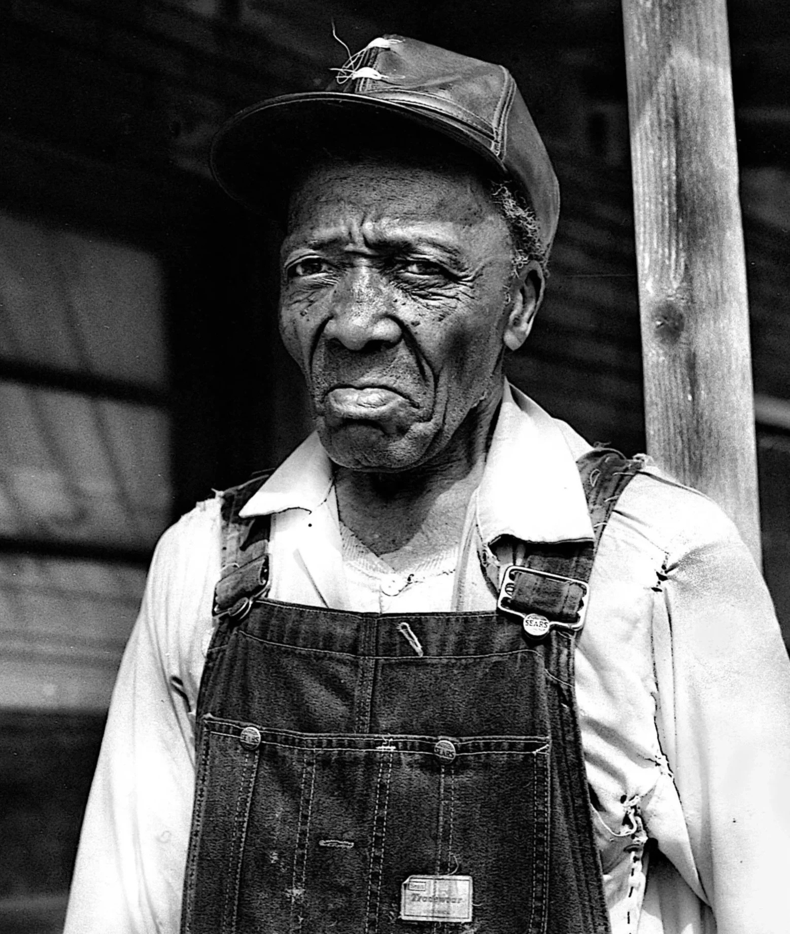 Black and white photo of an elderly man with deep wrinkles, wearing a cap and denim overalls, standing beside a wooden post.