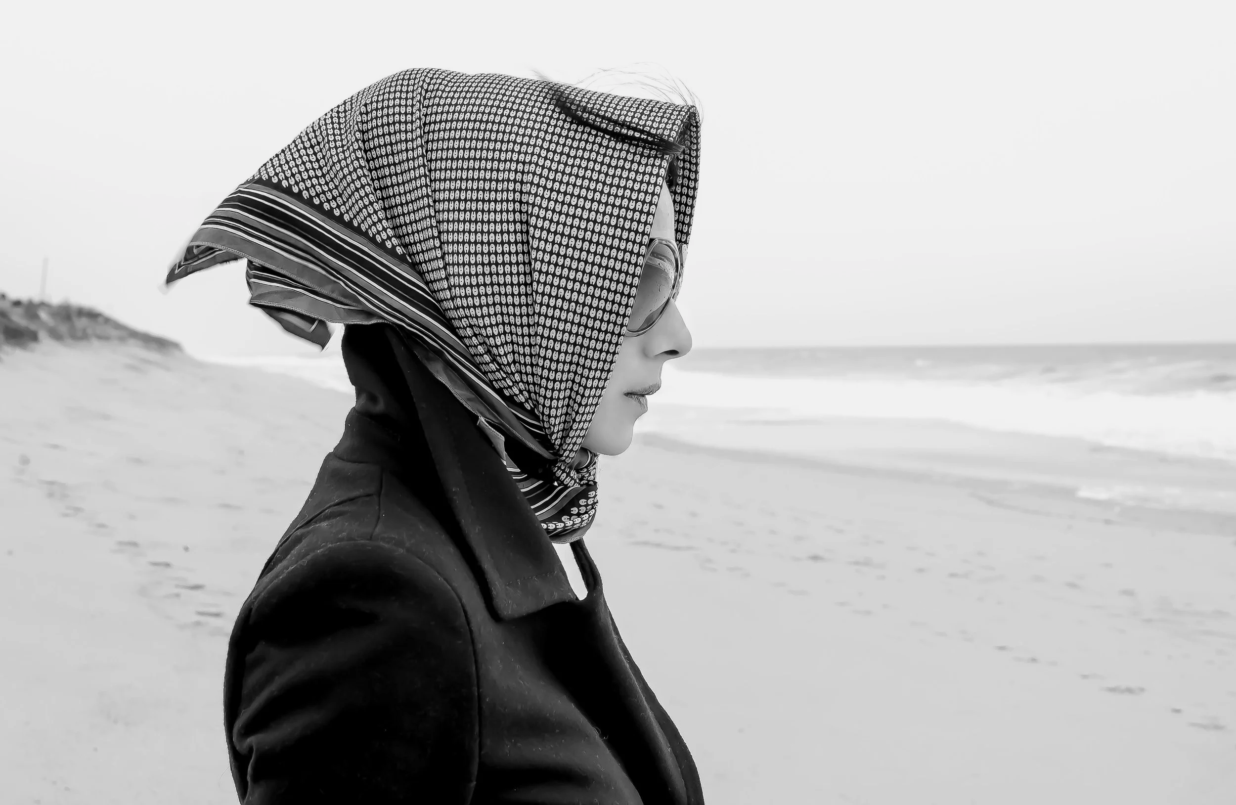A woman in profile wearing a headscarf and sunglasses stands on a sandy beach with the ocean in the background.