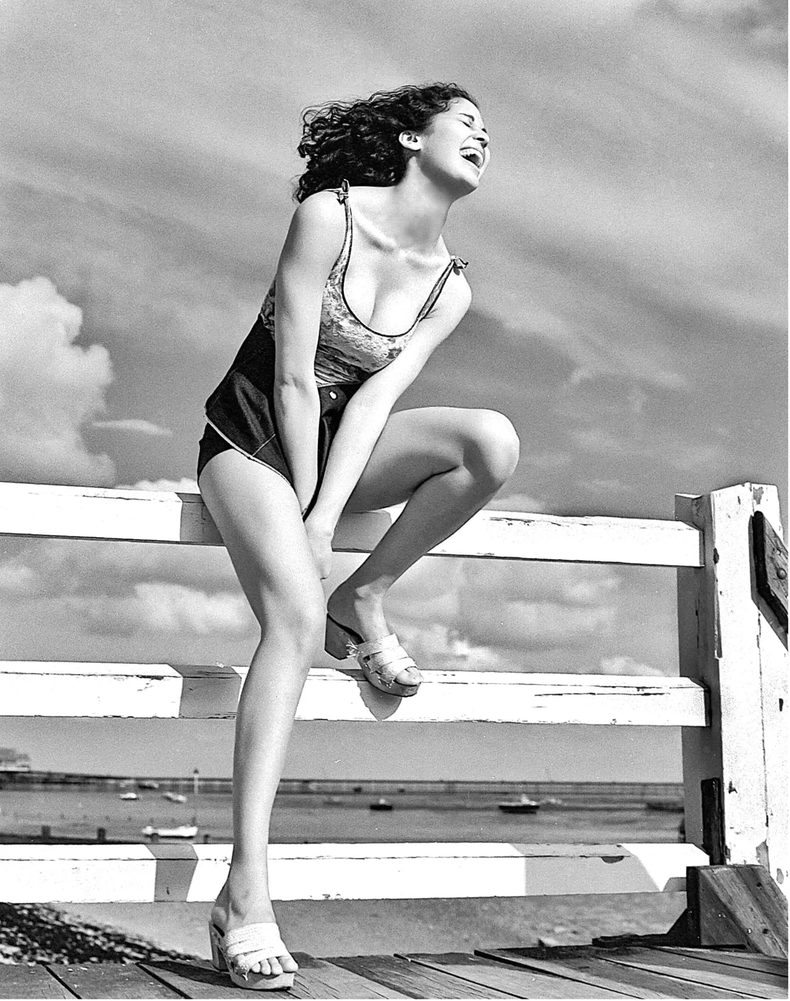 A woman with curly hair laughing, sitting on a wooden bench outdoors, wearing a tank top, shorts, and high-heeled shoes.