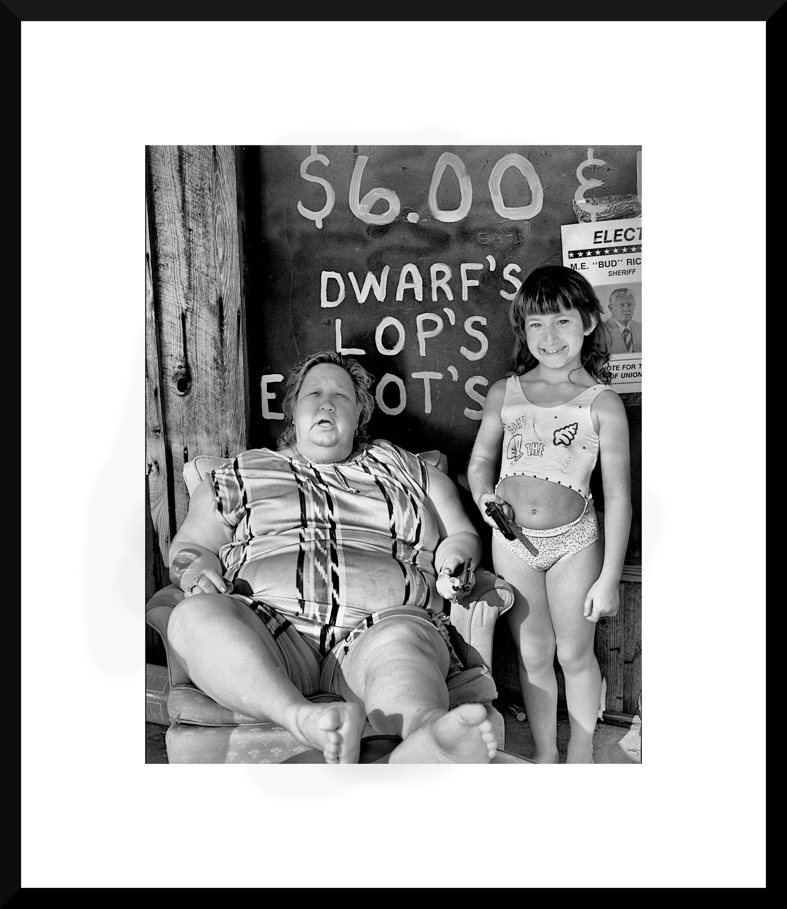 Black and white photograph of a woman sitting in a chair with a child standing next to her, both smiling. The woman is wearing a striped shirt, and the child is in a tank top and shorts holding a toy gun. Behind them is a sign that reads '$6.00 DWARF'S LOP'S EOT'S' and a political poster.