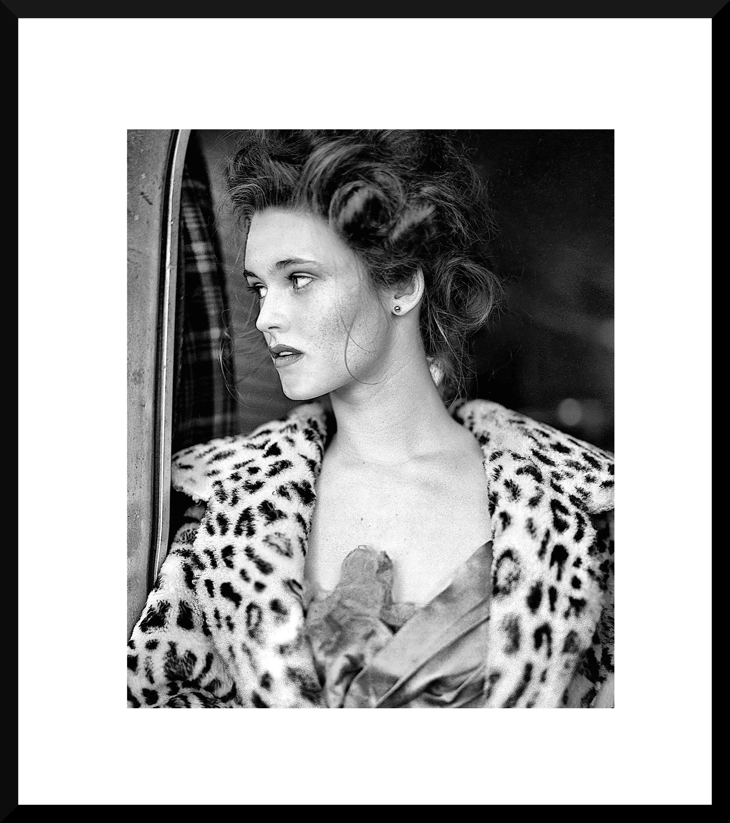 Black and white photograph of a woman with vintage style hair and makeup, wearing a leopard print coat, looking out a window with a contemplative expression.