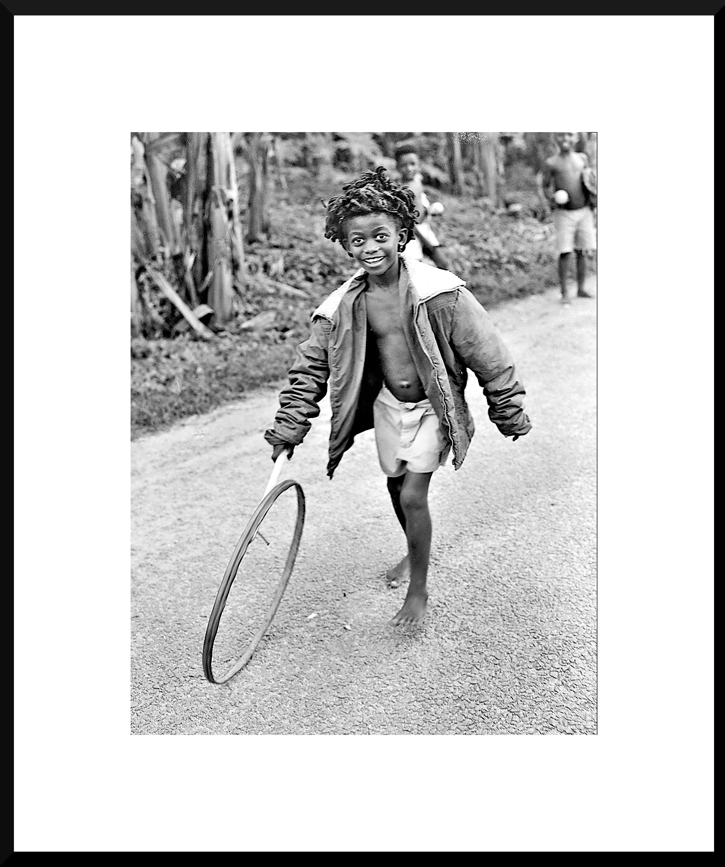 Black and white photo of a young boy running barefoot on a dirt road, holding a hula hoop, smiling, with other children in the background.