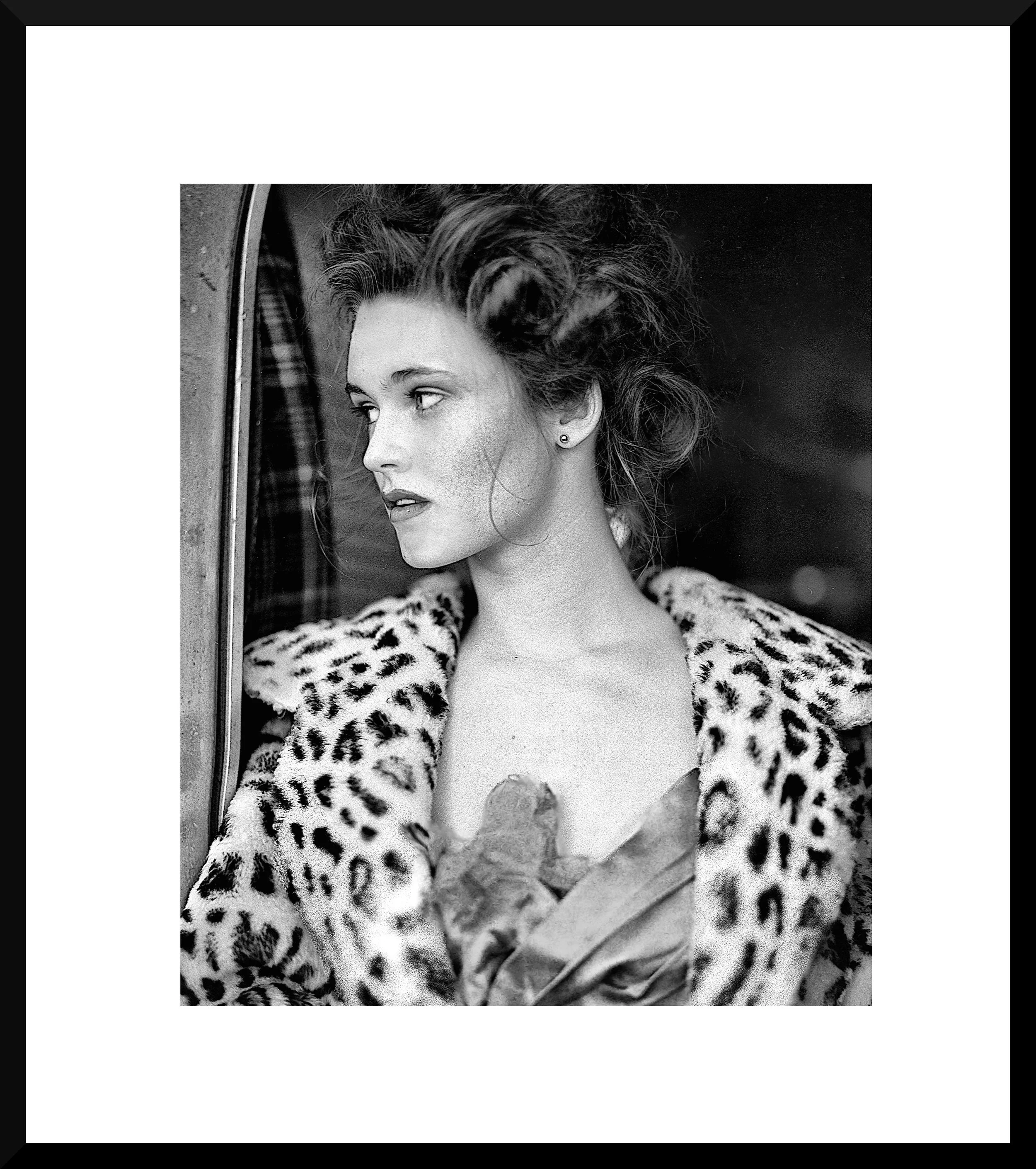 Black and white portrait of a woman with styled hair, wearing a leopard print coat, looking out of a window.