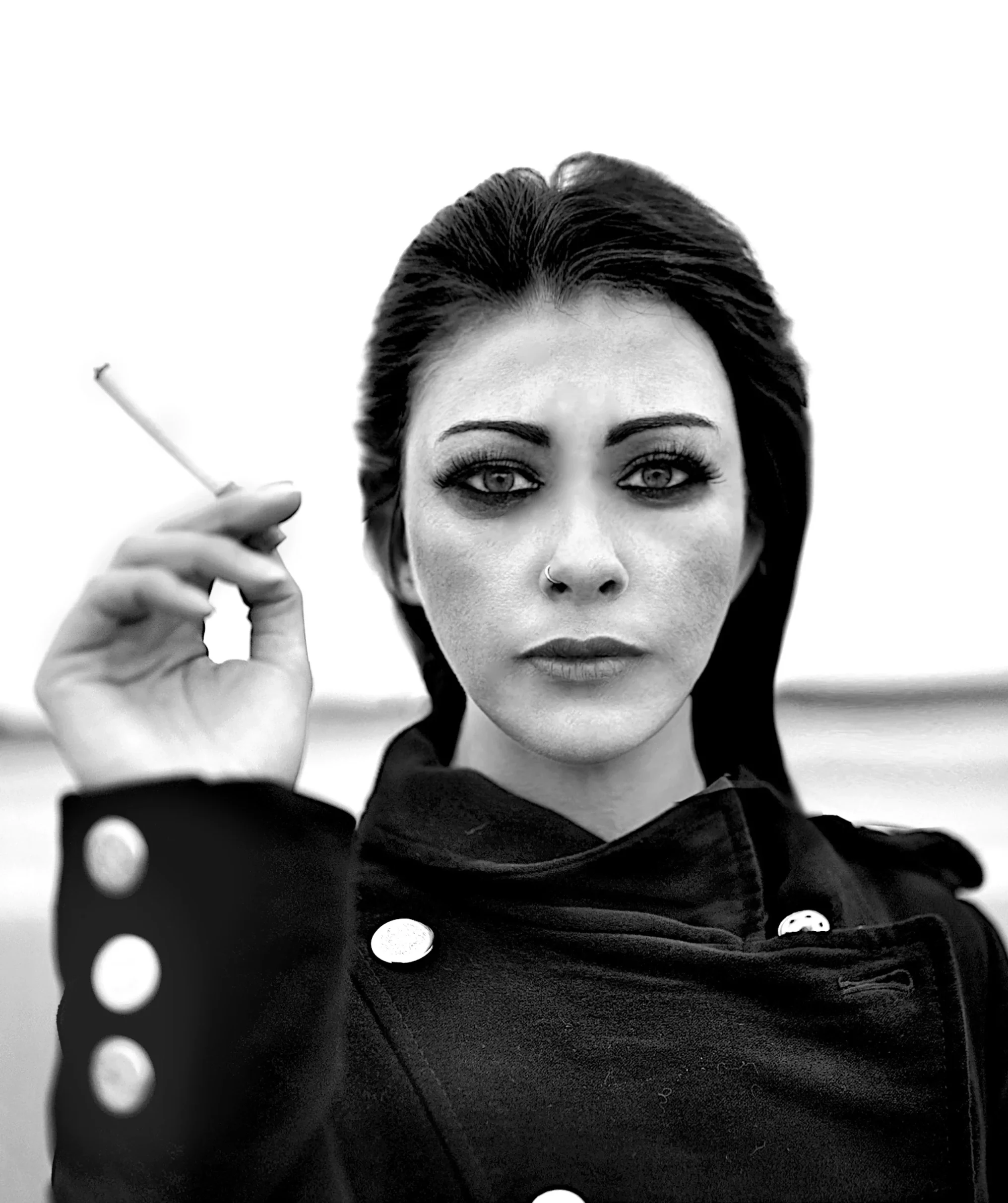 A woman with dark hair and makeup, wearing a dark coat, holds a cigarette near her face. The image is in black and white with a blurred background.