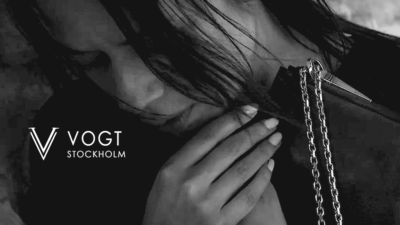 Close-up of a woman with long hair touching her face, with a chain and a strap nearby, and a logo that says "V VOGT STOCKHOLM" in the bottom left corner.