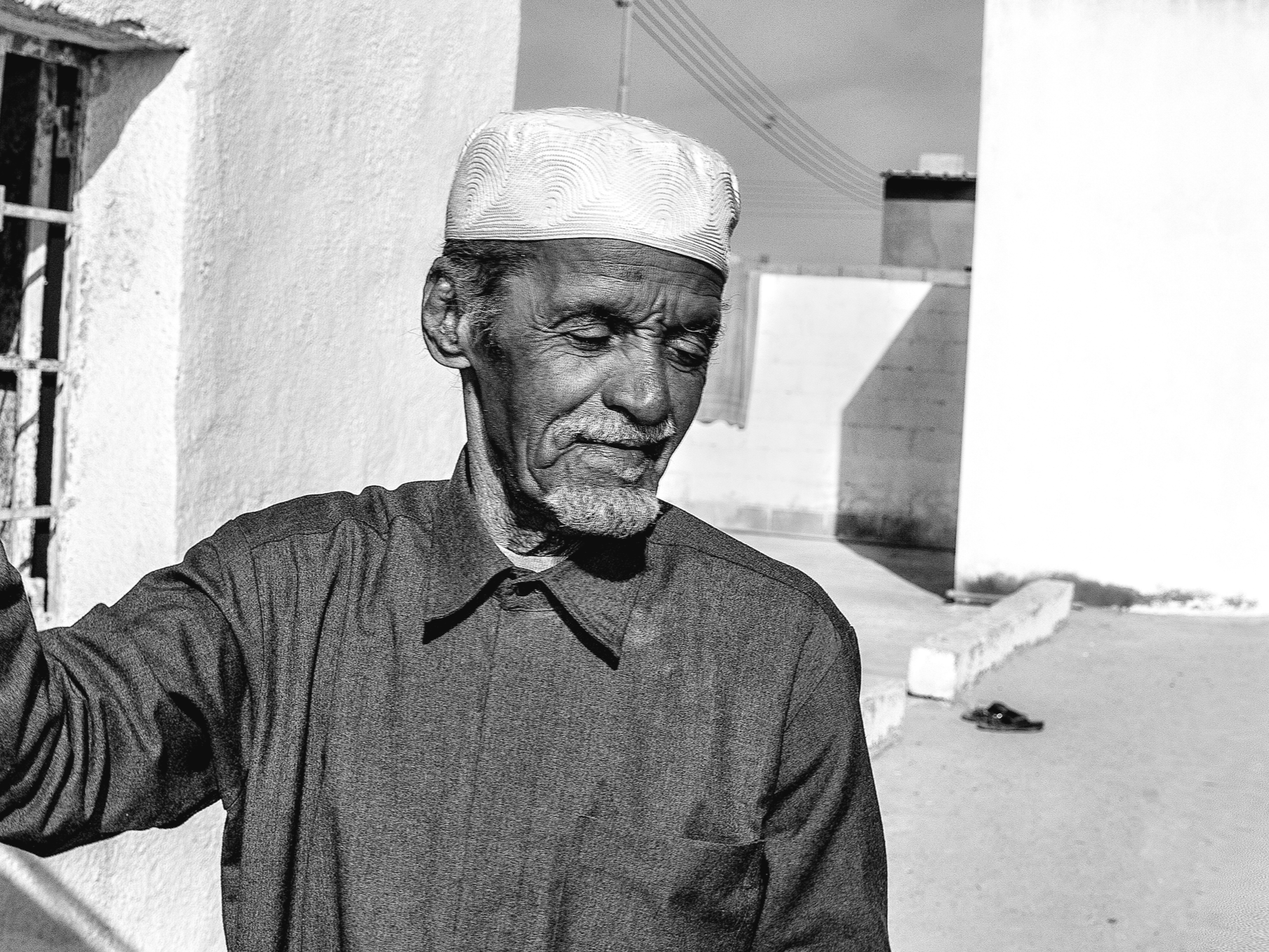 An elderly man with closed eyes, wearing a traditional cap and dark sweater, standing outdoors next to a white wall and a building, with sunlight casting shadows.