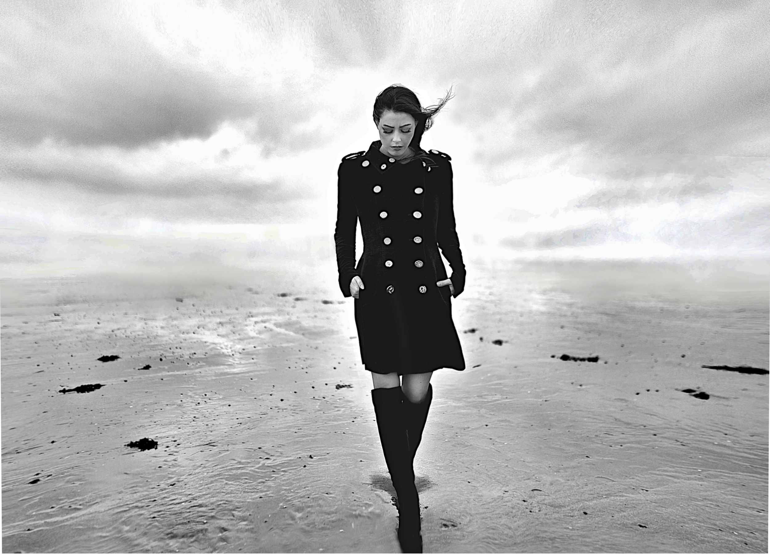A woman wearing a long black coat and high boots walking on a beach under cloudy skies.