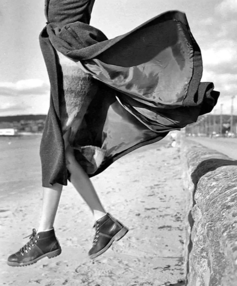 Person in mid-air jump at the beach, wearing dark boots, with a cloth or jacket flying around, and a stone wall in the foreground.
