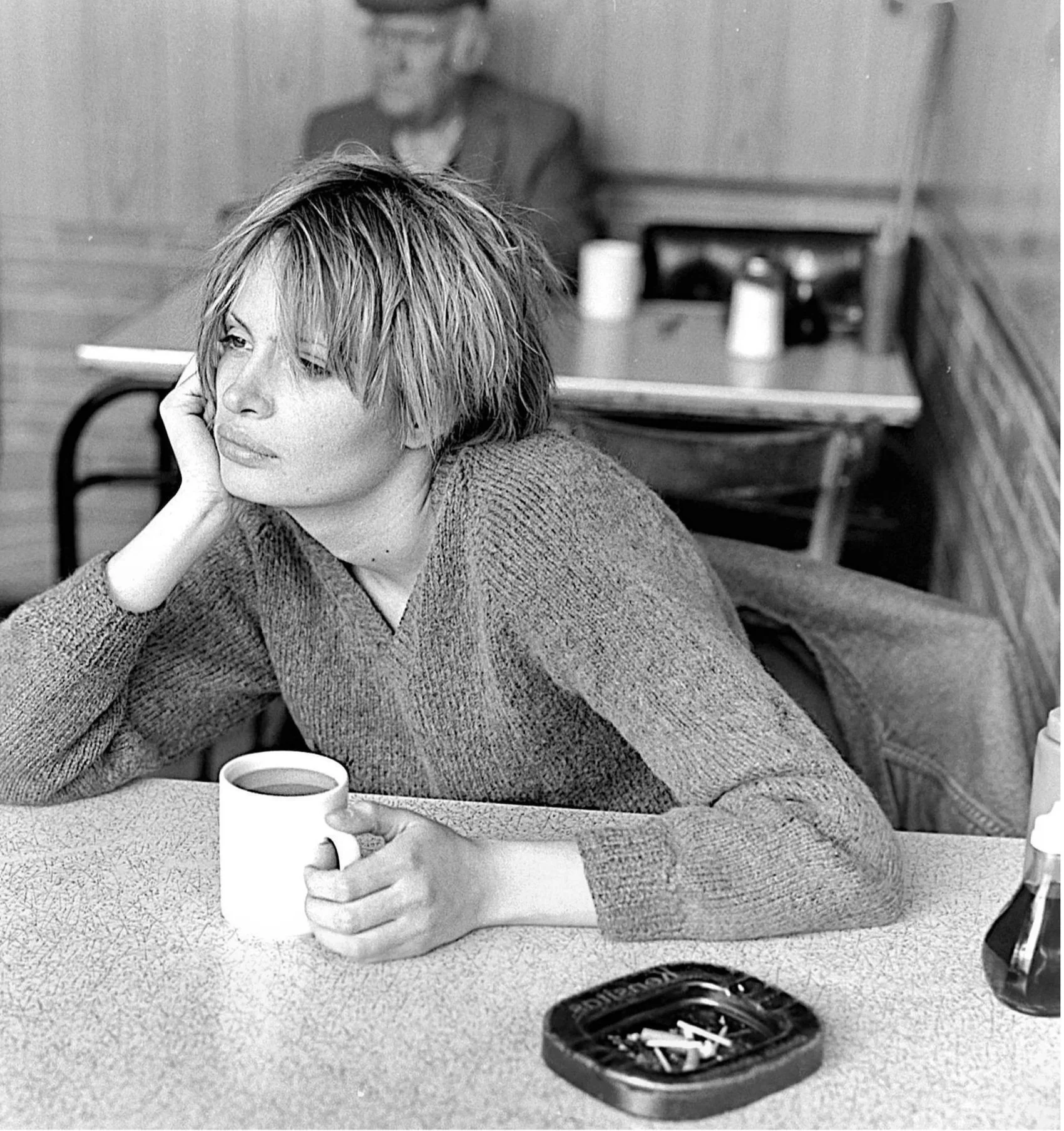 A woman with short, messy hair in a sweater sitting at a table, resting her head on her hand, holding a cup of coffee. In the background, a woman with glasses and a blazer sits at another table. There is an ashtray with cigarette butts on the table in the foreground.