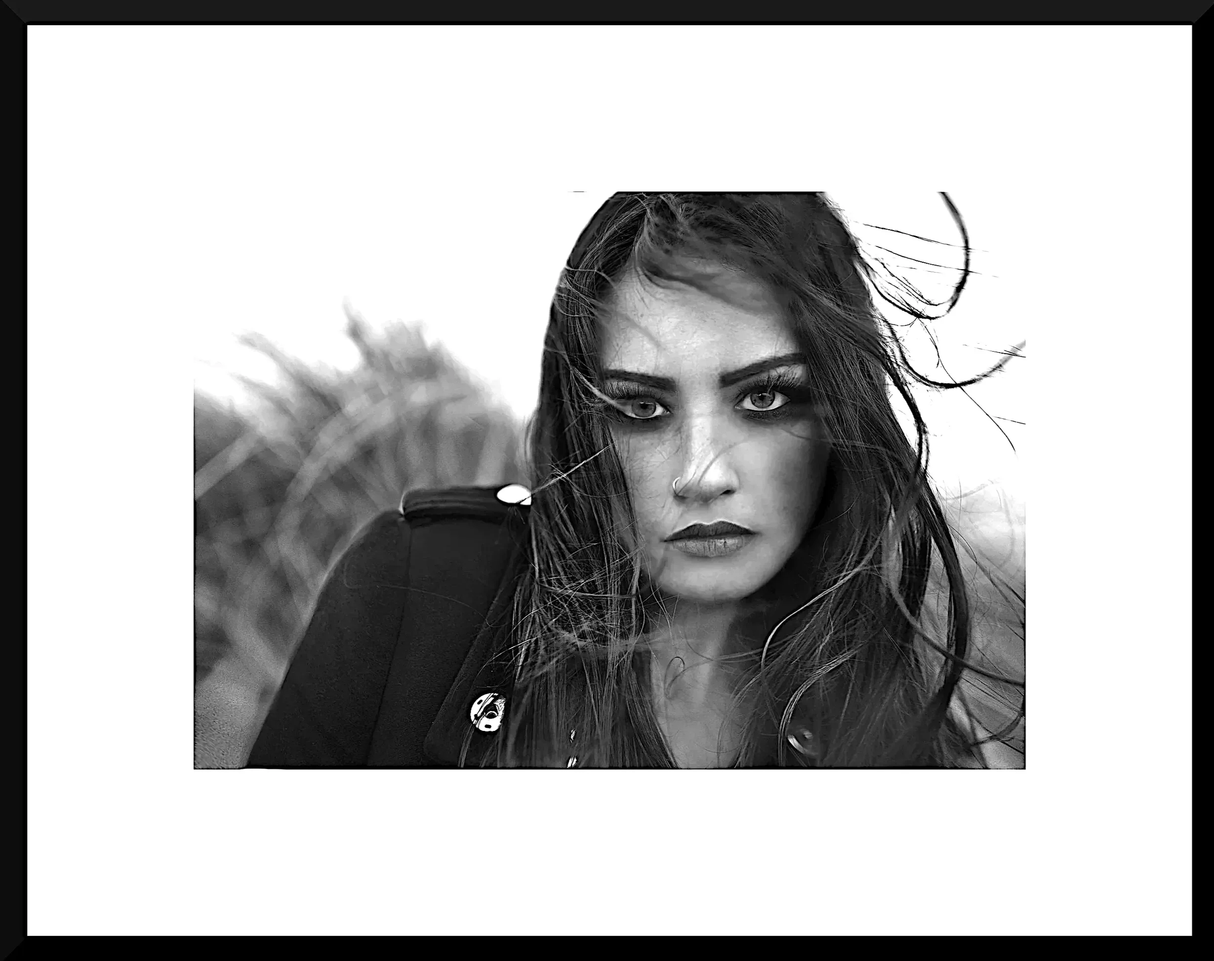 Black-and-white portrait of a woman with long, windswept hair, intense gaze, and bold makeup, outdoors with blurred nature background.