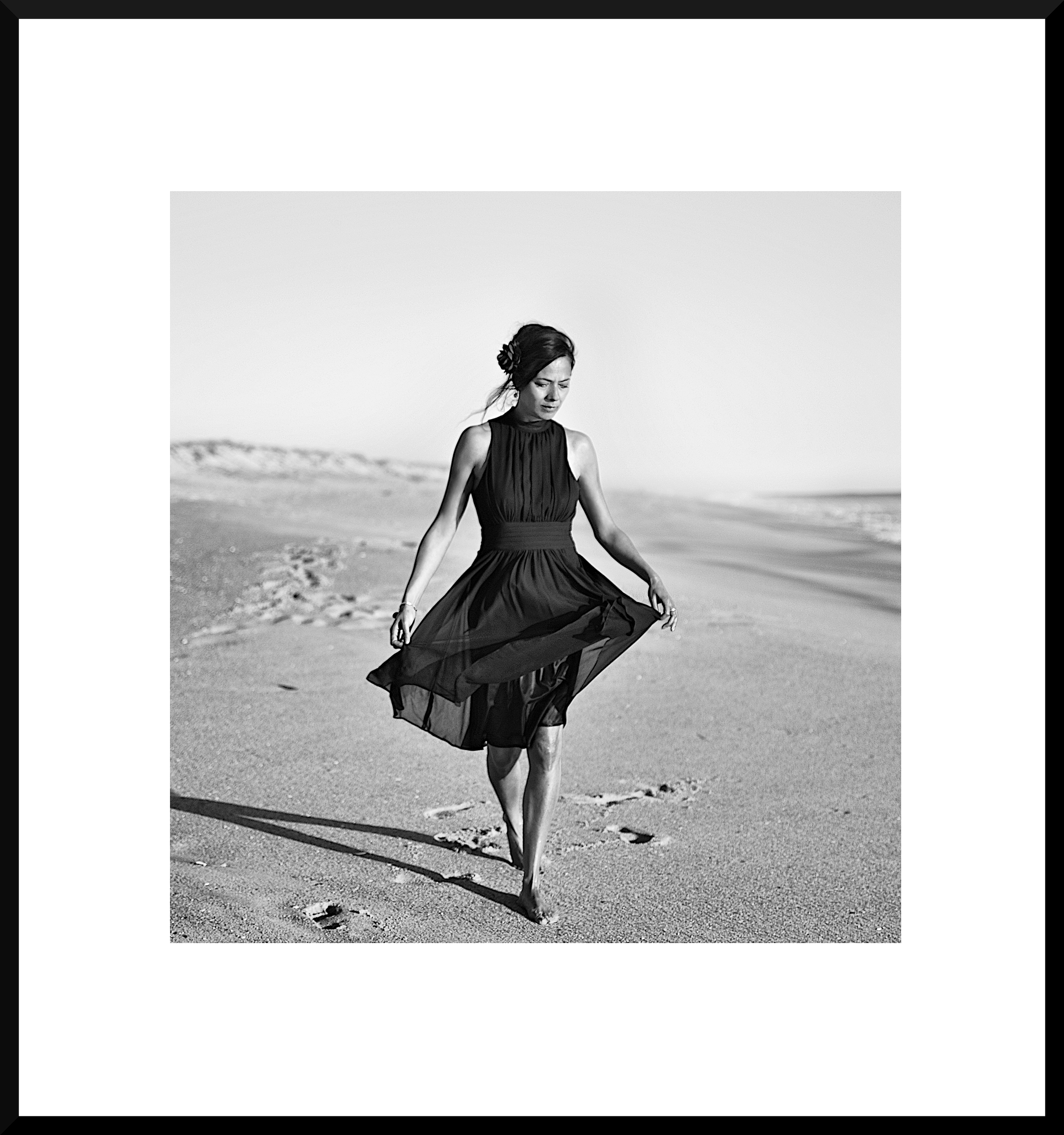 Black and white photo of a woman walking barefoot on the beach, wearing a dark sleeveless dress, holding the hem of her dress, with sand and ocean in the background.