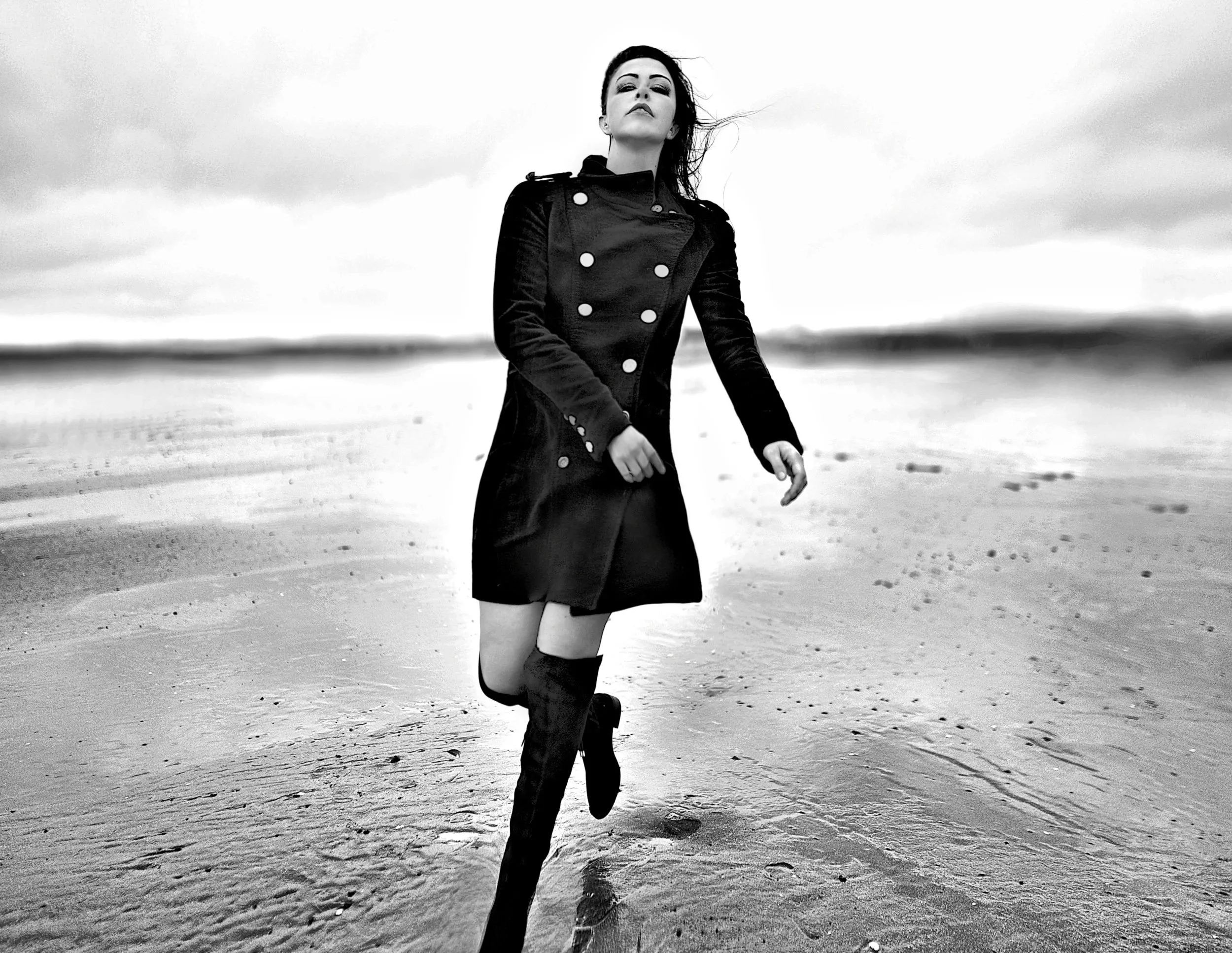 A woman wearing a black coat and knee-high boots running on a sandy beach during daytime with clouds in the sky.