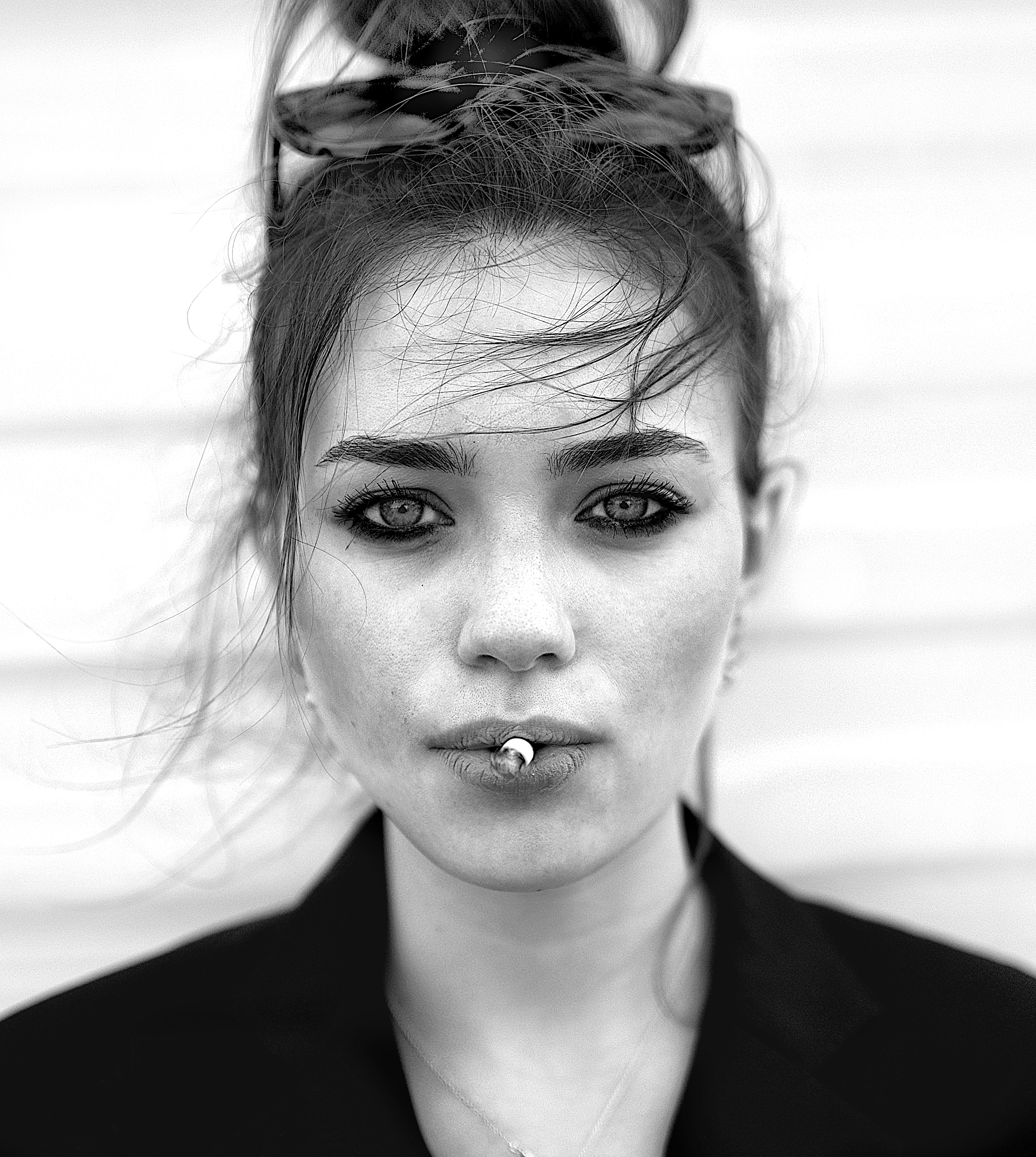 Close-up black and white portrait of a woman with striking eyes, smoky eye makeup, and slightly messy hair styled in an updo with a large bow, wearing a black outfit with a deep neckline.