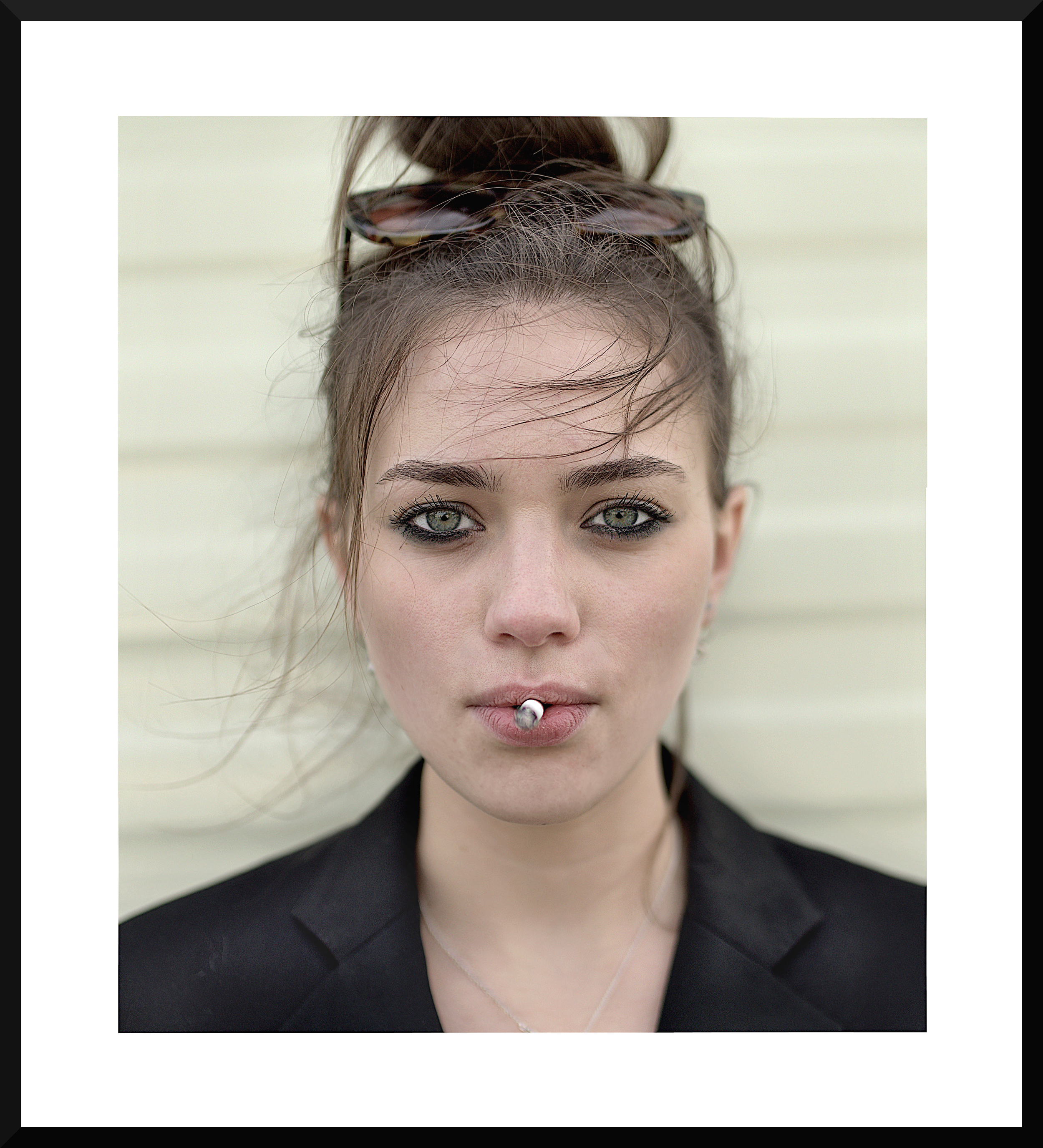 A close-up portrait of a woman with blue eyes and light skin, wearing a black jacket and sunglasses on her head, with slightly messy hair and a cigarette in her mouth.