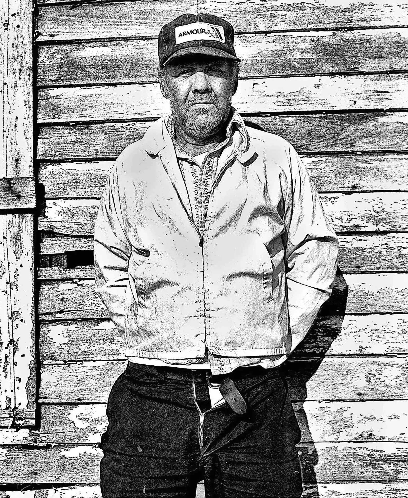 Black and white photo of an older man standing in front of a wooden wall. He is wearing a cap with the words 'ARMOUR' on it, a zip-up jacket, a button-up shirt, and dark pants.