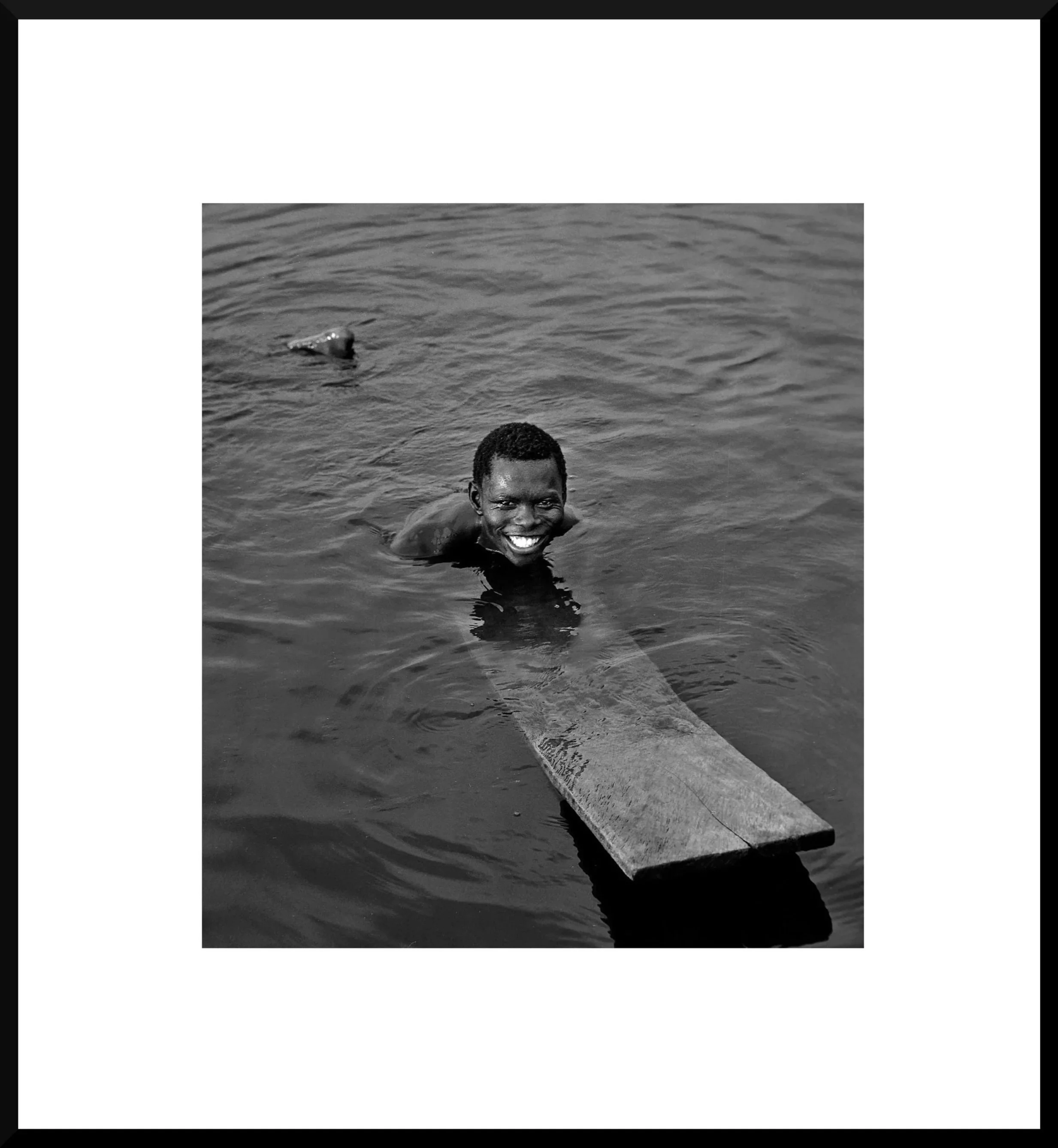 A smiling person swimming in a body of water with a piece of wood or plank, with bank or shoreline in the background.