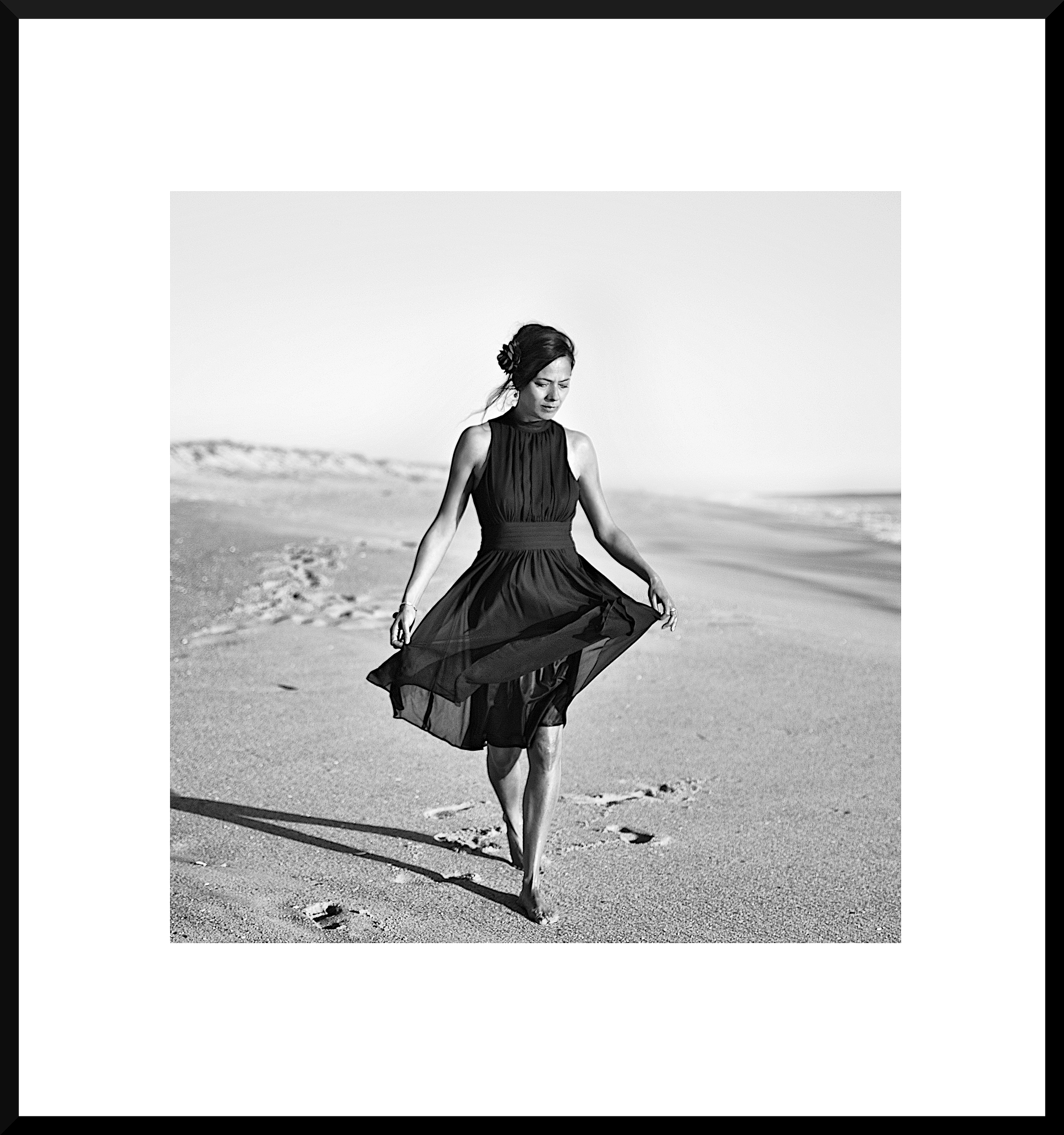 A woman in a black dress walking barefoot on the beach, holding the hem of her dress, with footprints in the sand, in black and white.