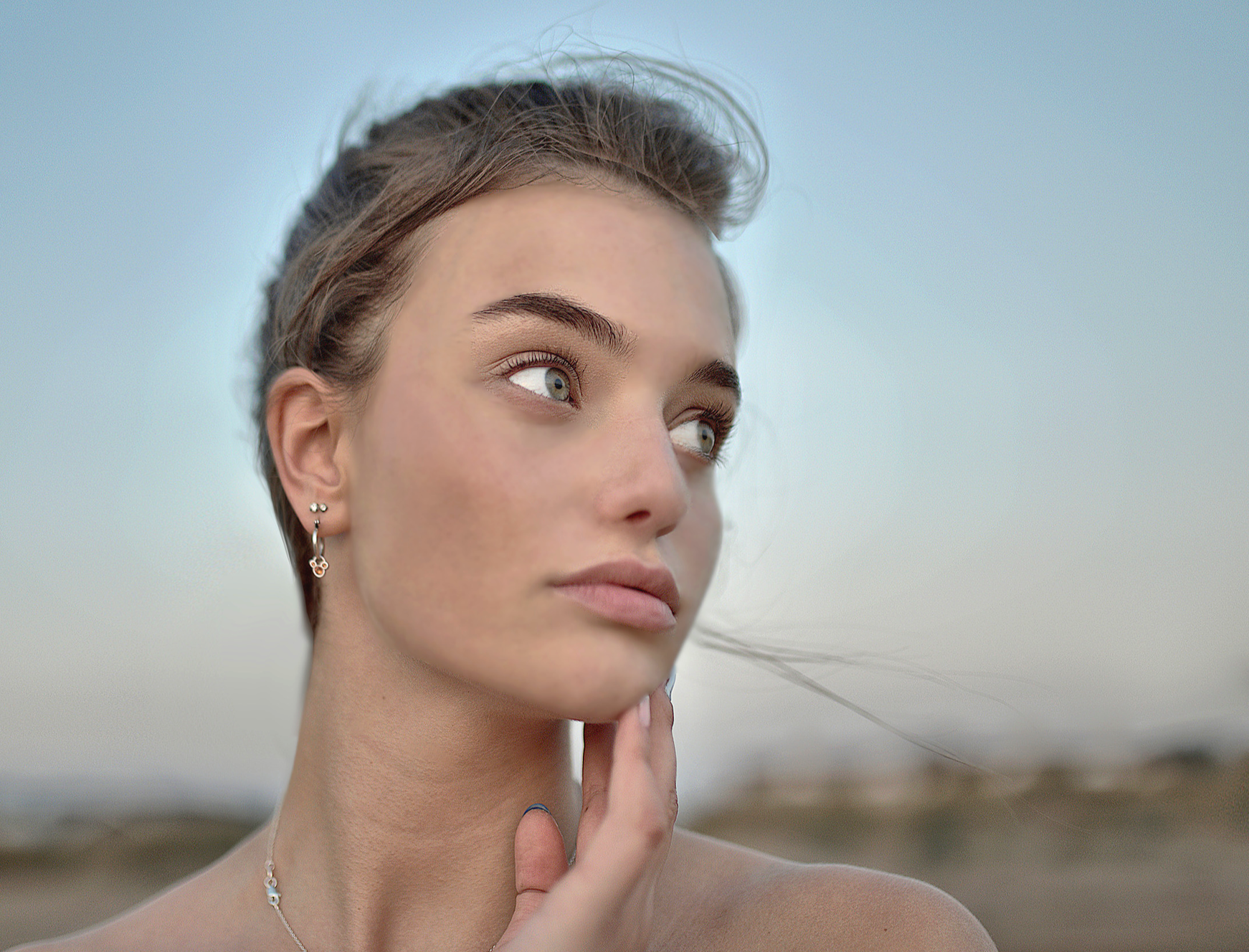 Close-up of a young woman with light brown hair, wearing earrings and a necklace, gazing thoughtfully into the distance with her hand near her chin against a soft, blurred background.