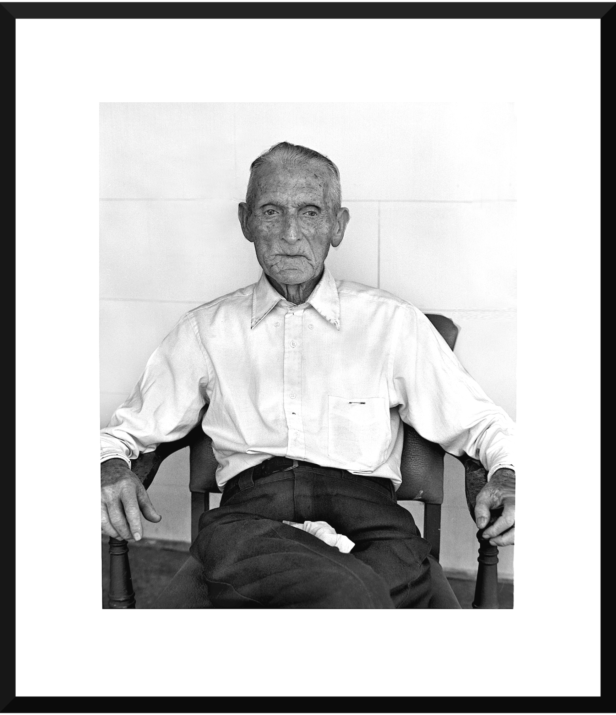 Black and white photo of an elderly man sitting in a chair, wearing a collared shirt and dark pants, looking directly at the camera with a serious expression.