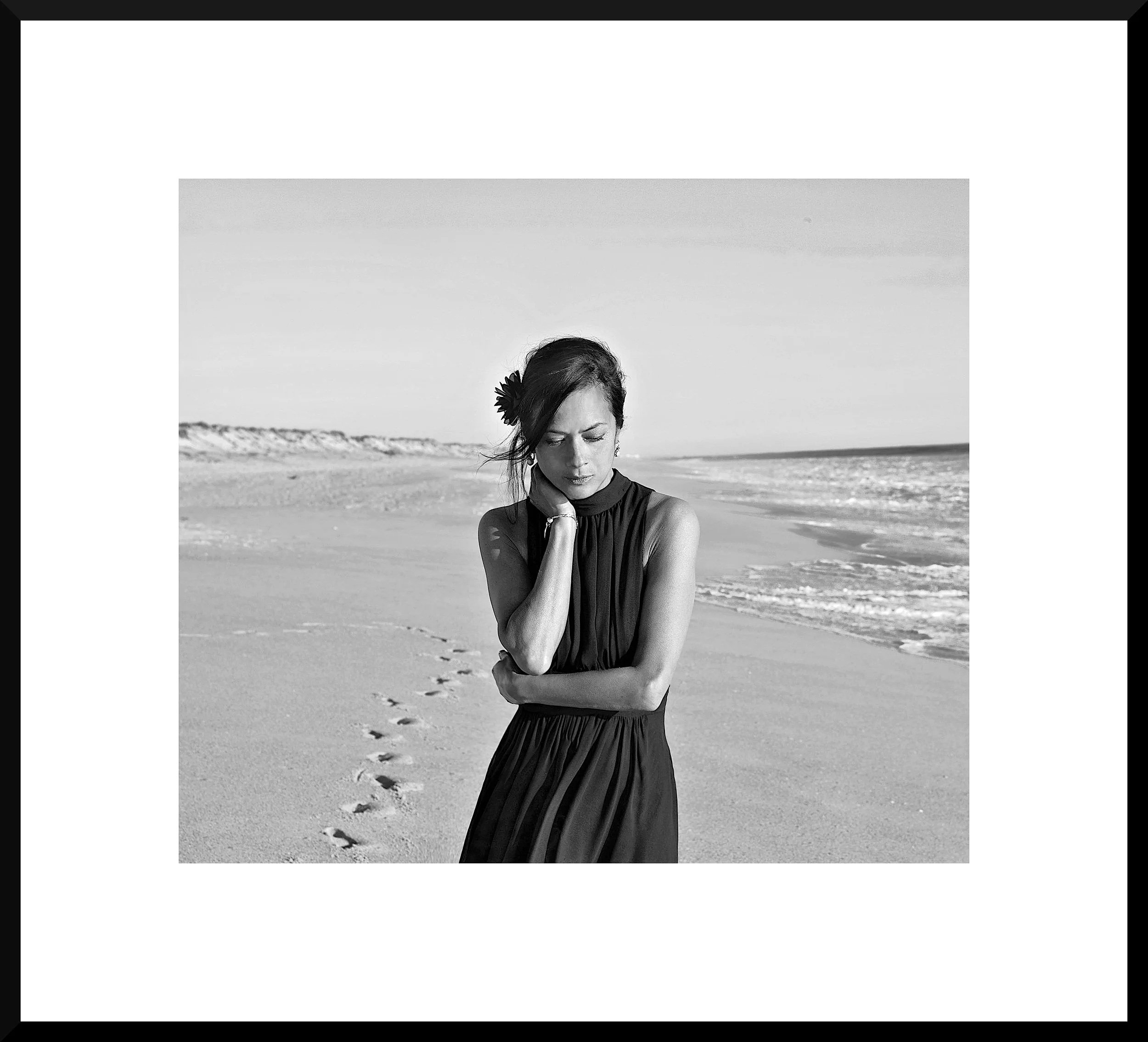 Black and white photograph of a woman standing on a beach, looking down with her hand touching her neck, footprints in the sand, ocean waves in the background.