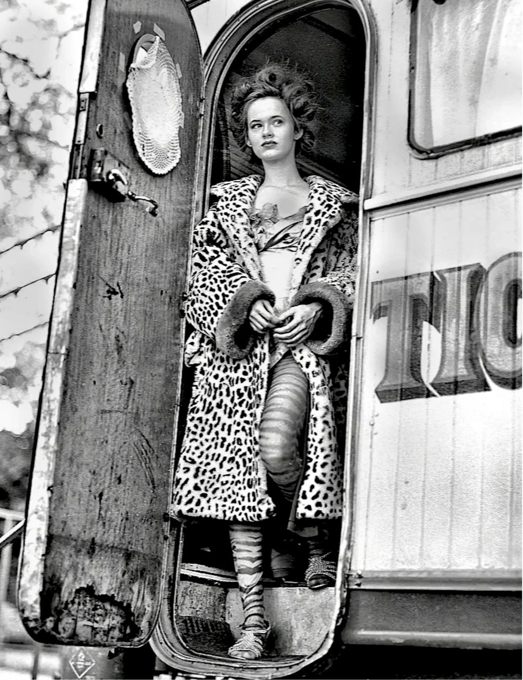 A woman with curly hair wearing a leopard print coat and striped stockings, standing in a rounded opening of a caravan or camper with her hands clasped in front of her. Her expression is neutral. Behind her, a large animal's head, possibly a lion, is visible, blending into the background.