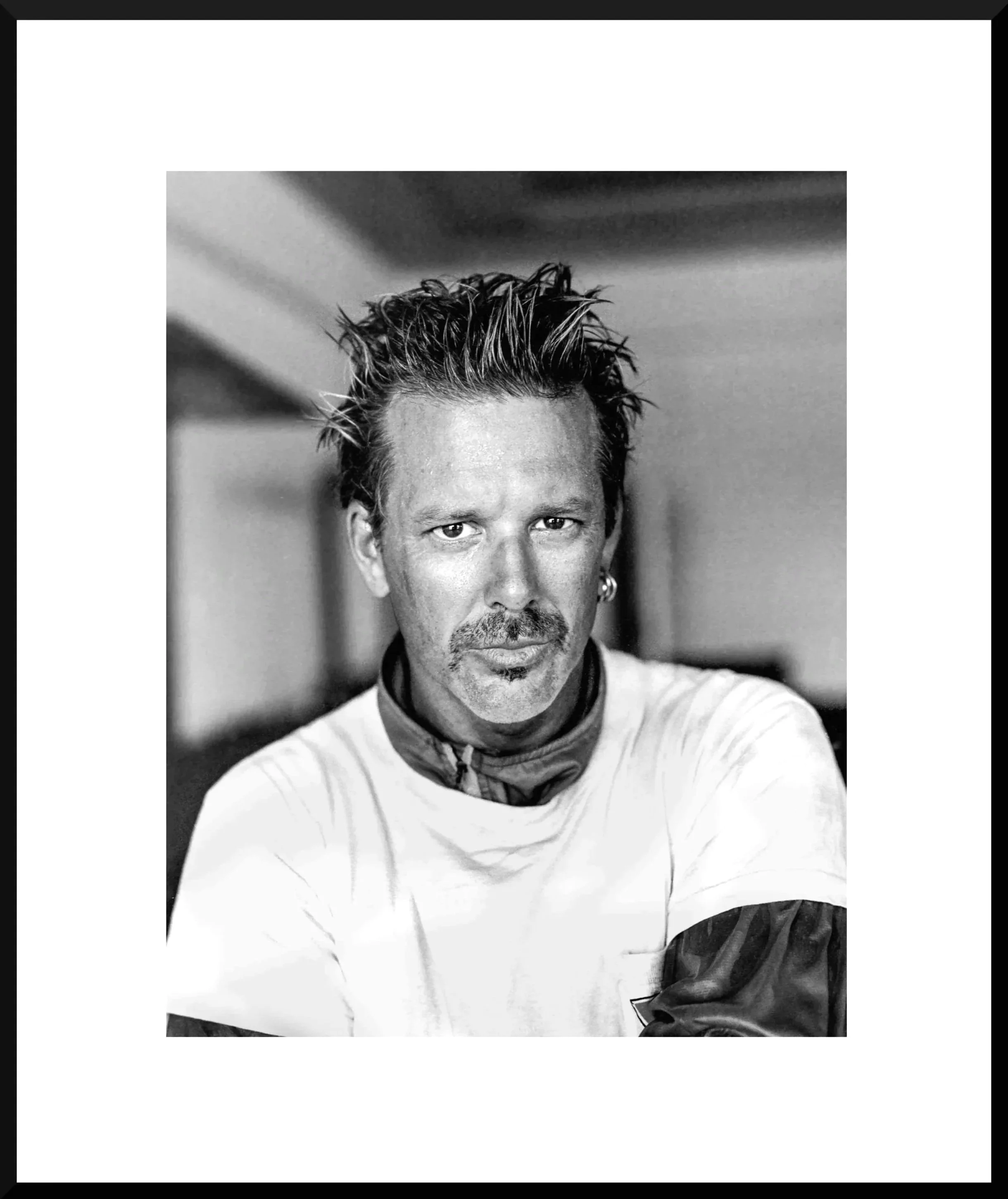 Black and white portrait of a man with spiky hair, a mustache, and a beard, wearing a white shirt and a patterned shirt underneath, looking directly at the camera.