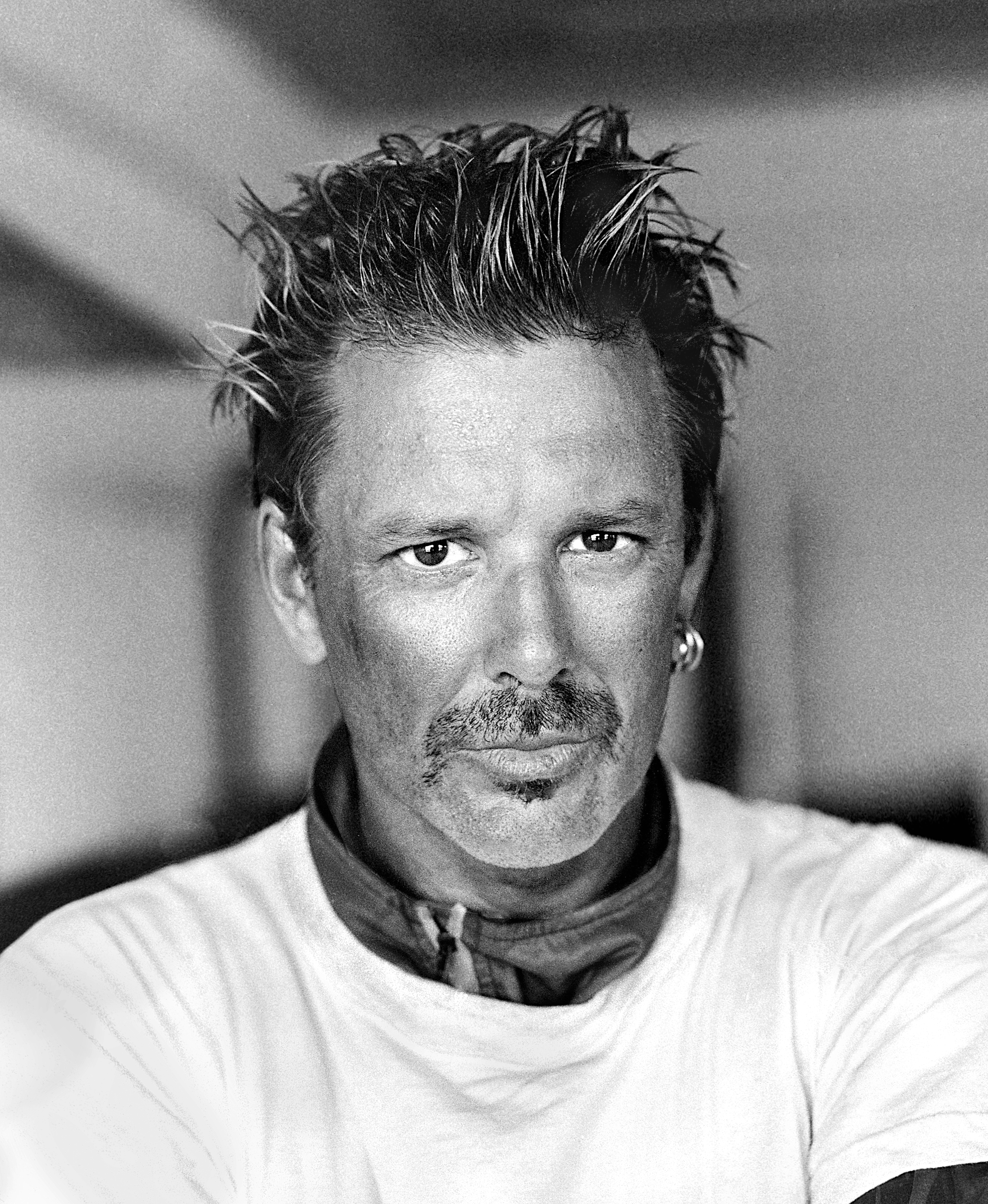 Black and white portrait of a man with spiked hair, a mustache, and goatee, wearing a light-colored shirt and earrings.