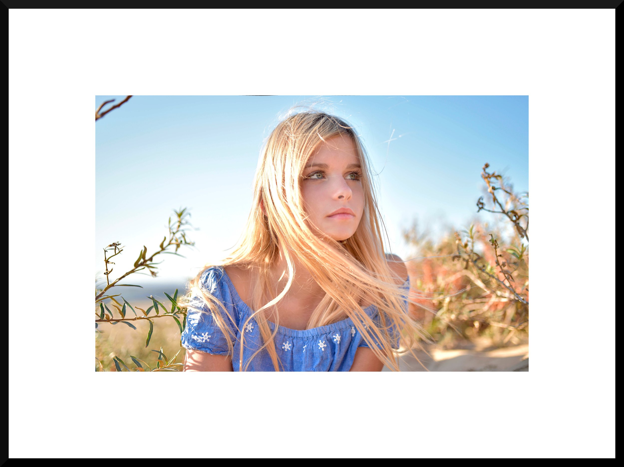 Young woman with long blonde hair wearing a blue dress, standing outdoors among bushes under a clear blue sky.