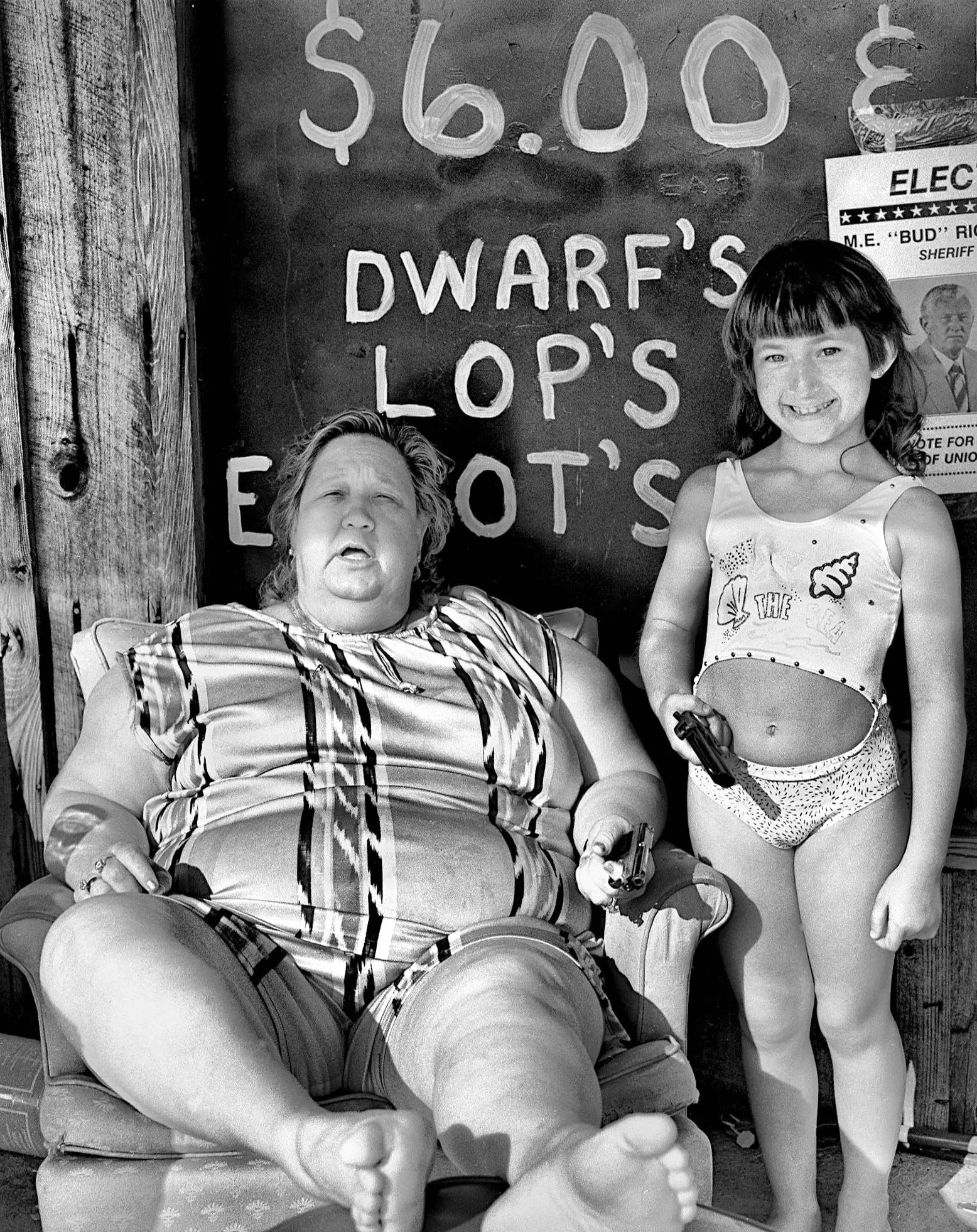 A woman with short hair and a young girl with long hair smiling outdoors next to a chalkboard with signs about food prices and election posters.