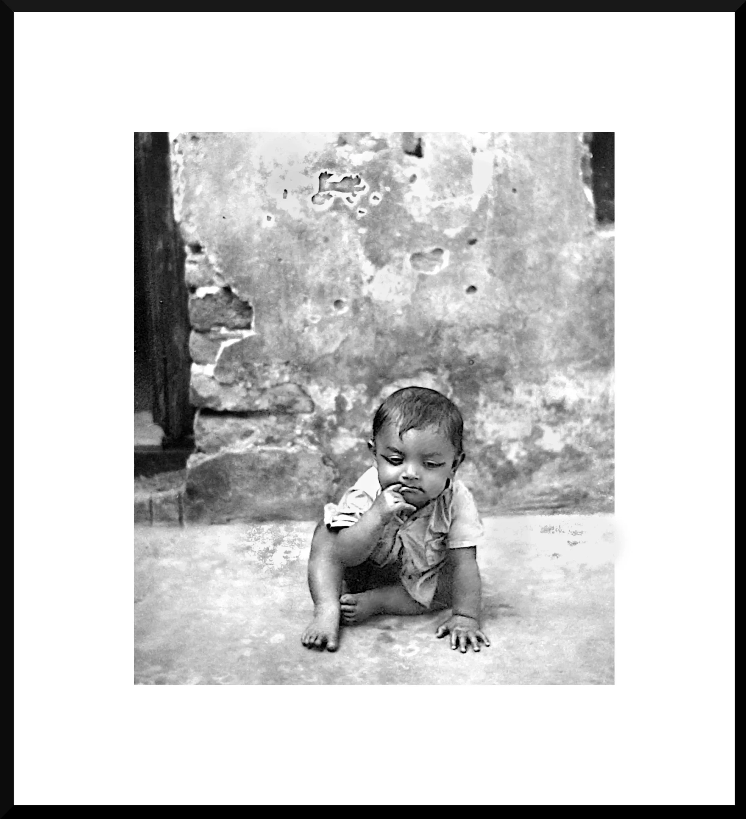 Black and white photo of a young child sitting on the ground with one hand near their mouth, in front of a weathered stone wall.