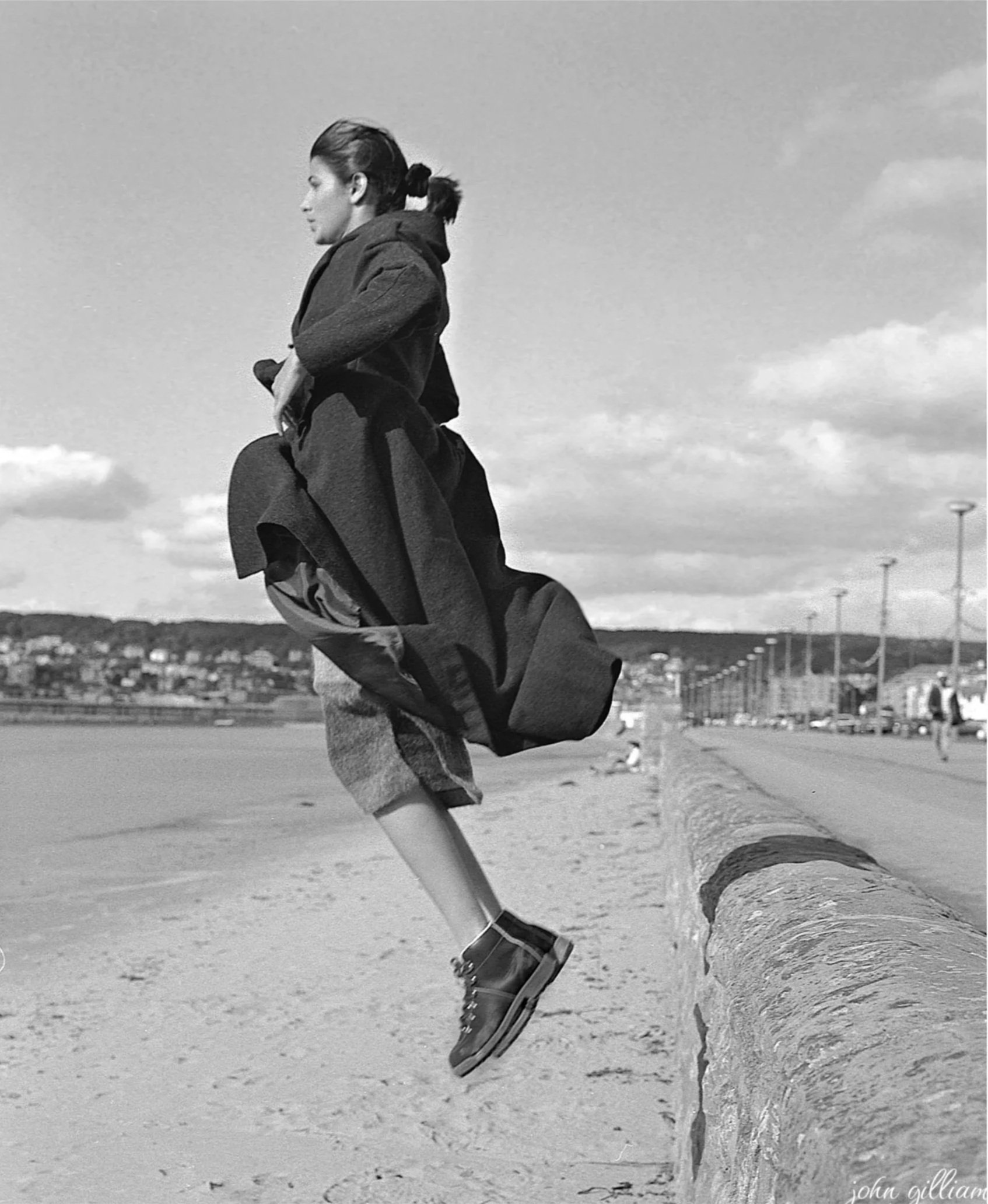 Black-and-white photo of a woman in a hoodie and jeans jumping high into the air with rollerblades on, in an outdoor setting with a cloudy sky and a cityscape in the background.