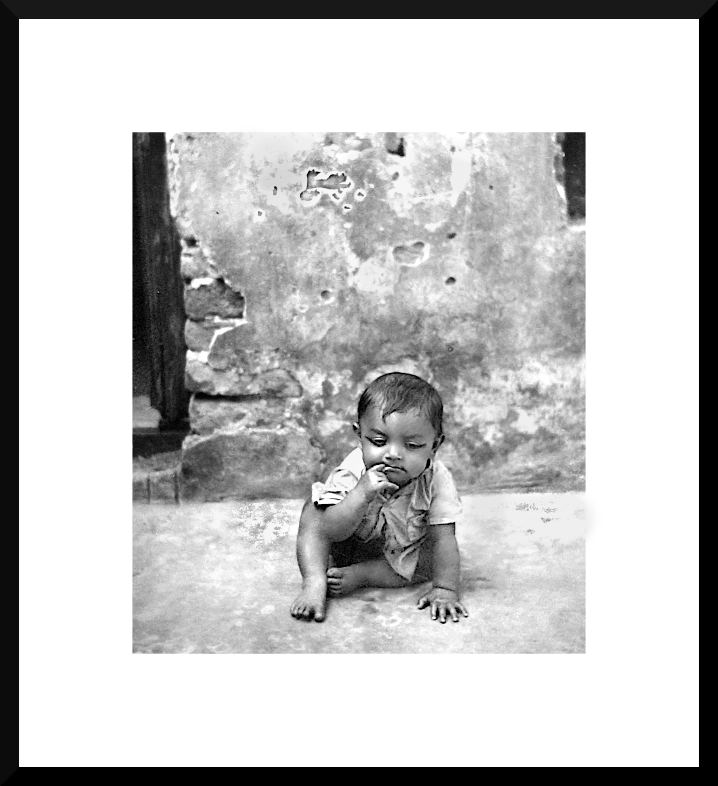Black and white photo of a young child sitting on the ground with one hand on his chin, against a worn, textured wall.
