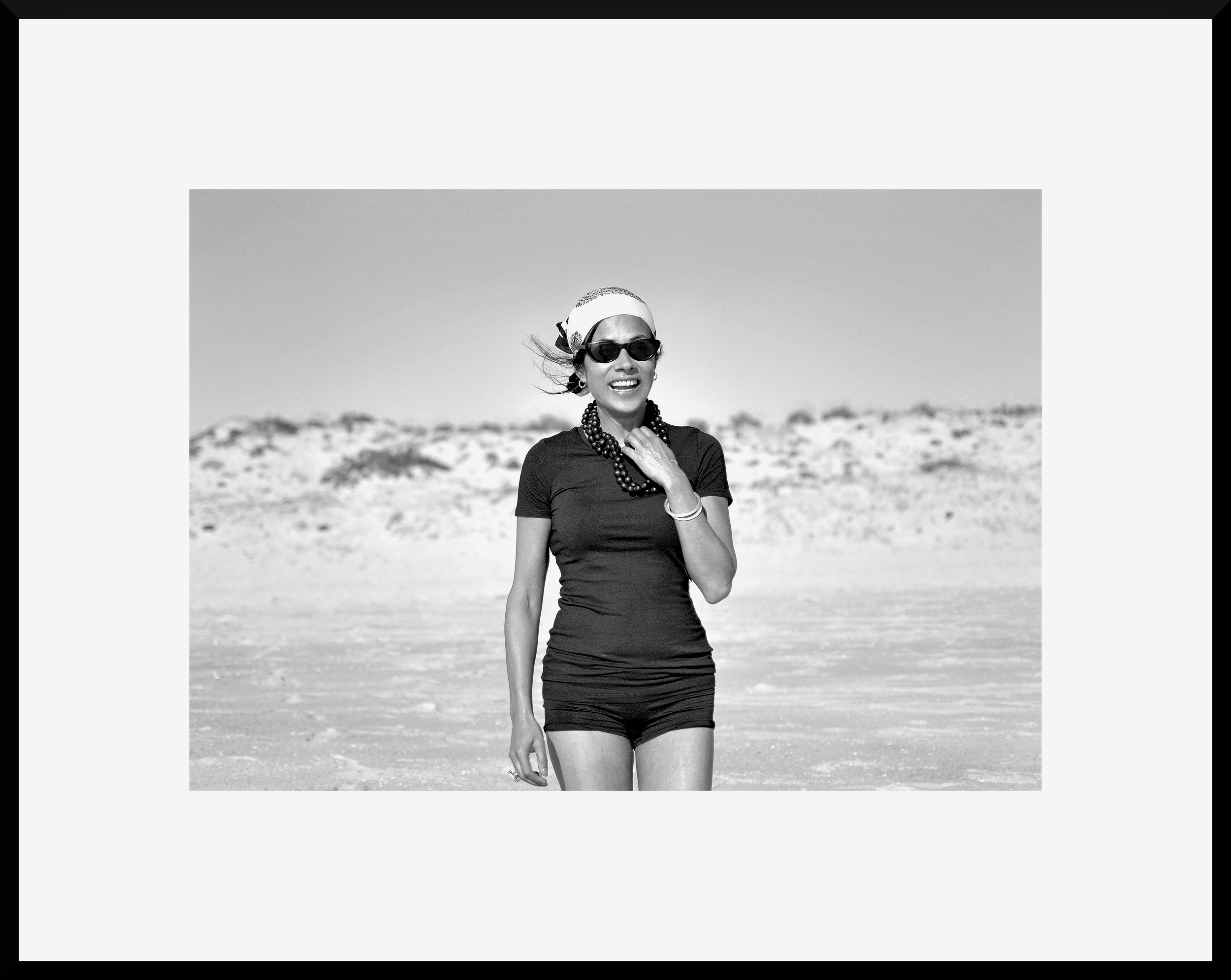 A woman smiling and walking on a beach in summer, wearing sunglasses, a headscarf, a necklace, a short-sleeved shirt, and shorts.